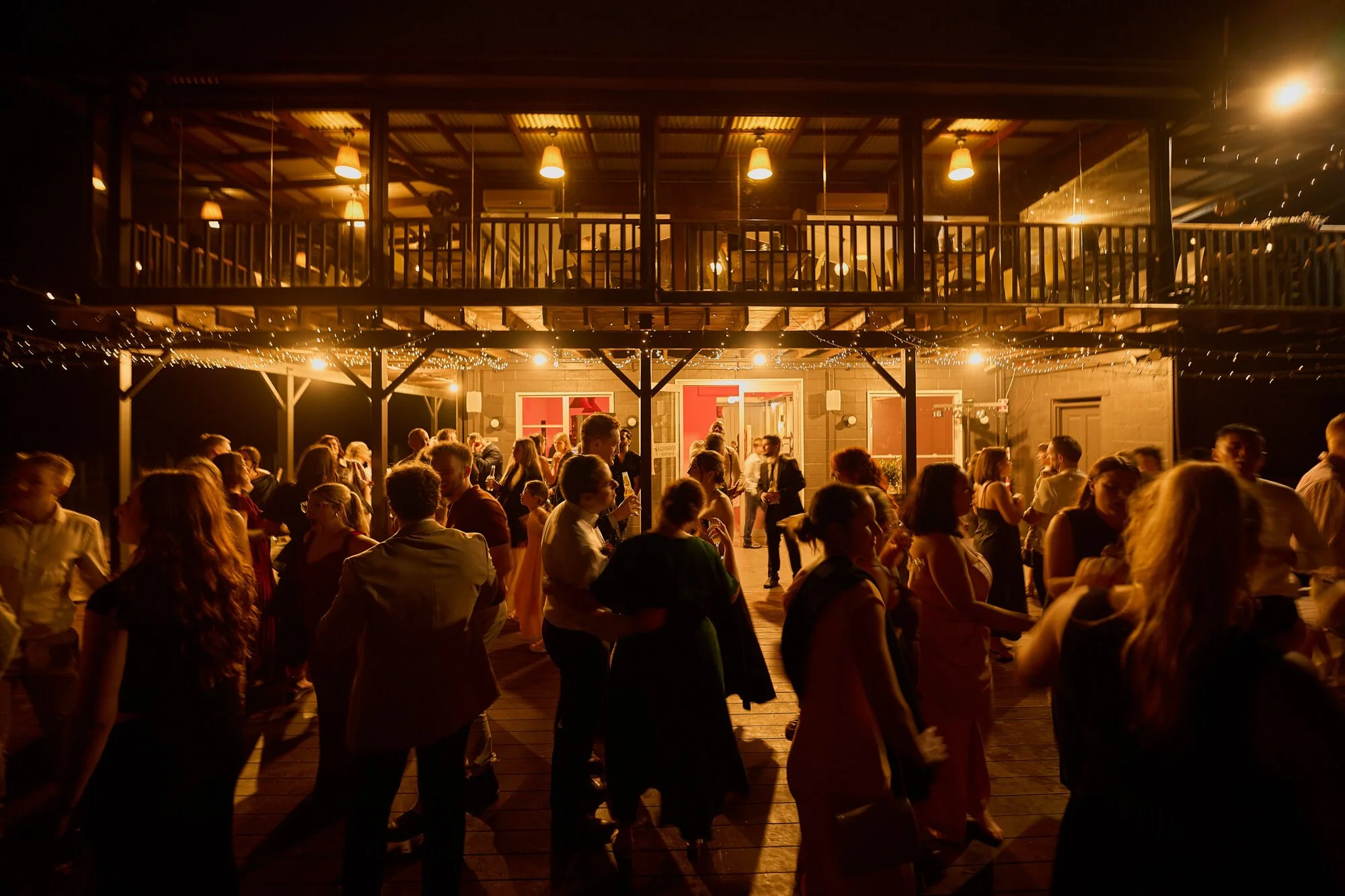 People dancing and socializing at an outdoor evening party with warm lighting, string lights, and a two-story building with a balcony in the background.