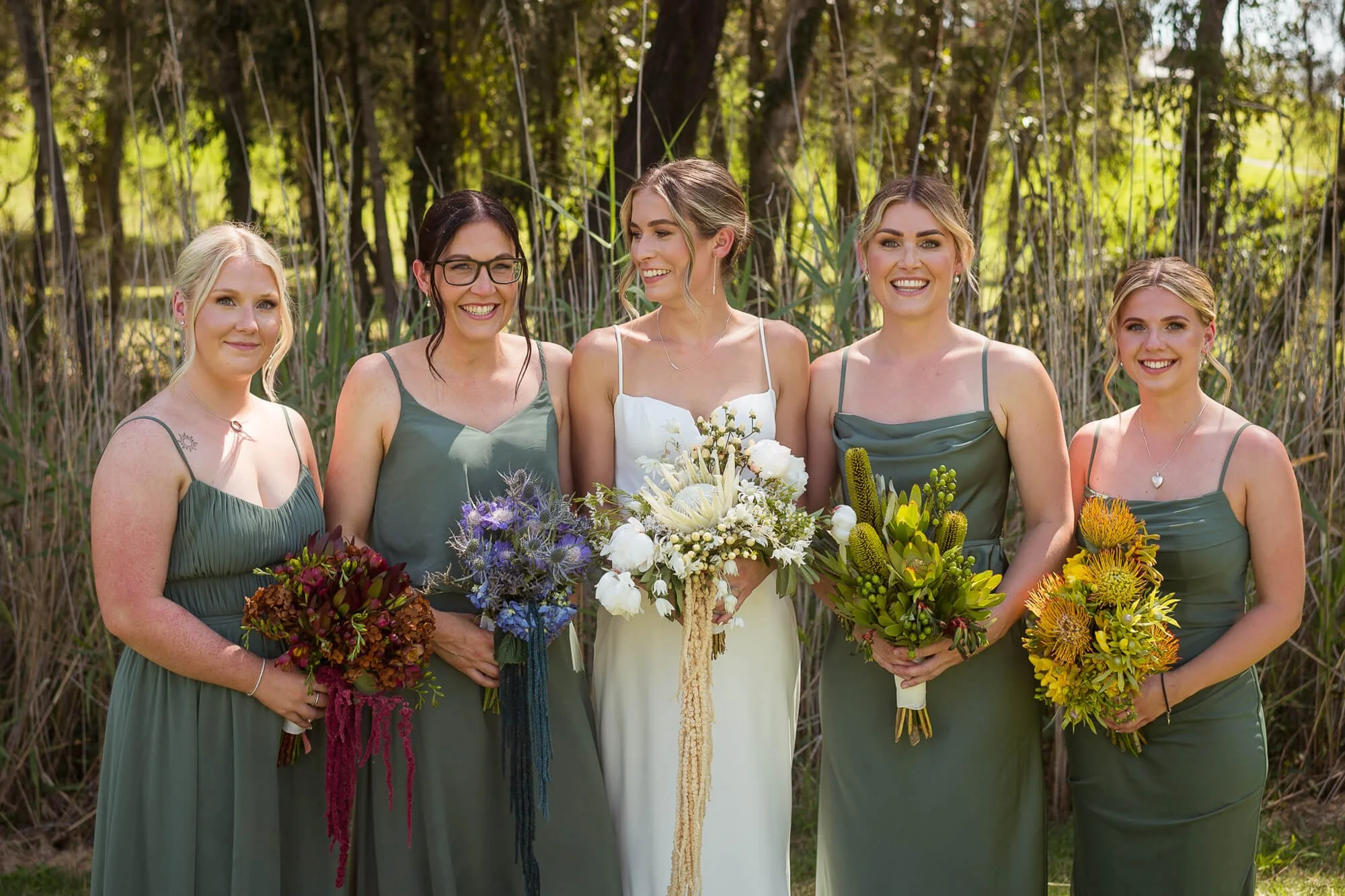 Group of five women in green dresses, one in white, at a wedding, holding bouquets of flowers, standing outdoors with trees in background.