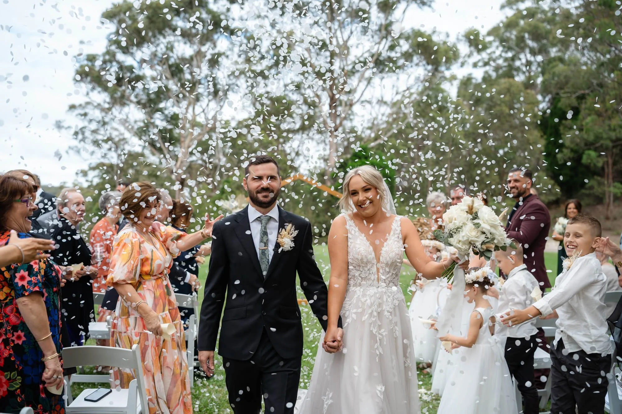 A newlywed couple walks hand in hand through a shower of confetti, surrounded by guests celebrating outdoors with trees in the background.