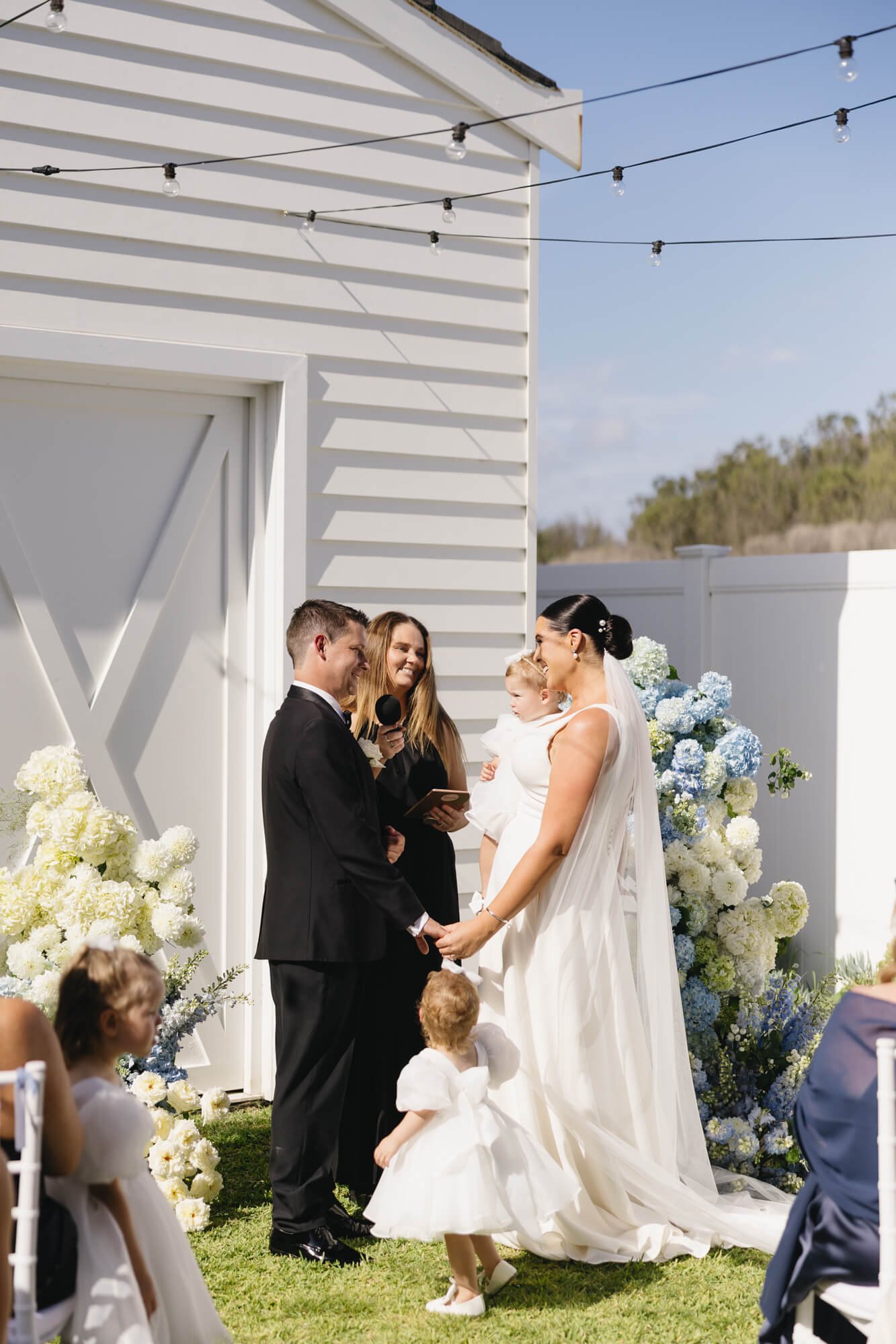 A wedding ceremony outdoors with a bride and groom holding hands, facing each other, during their vows. The bride is wearing a white dress and veil, and the groom is in a black suit. There are flowers and guests around them, and a person holding a mi