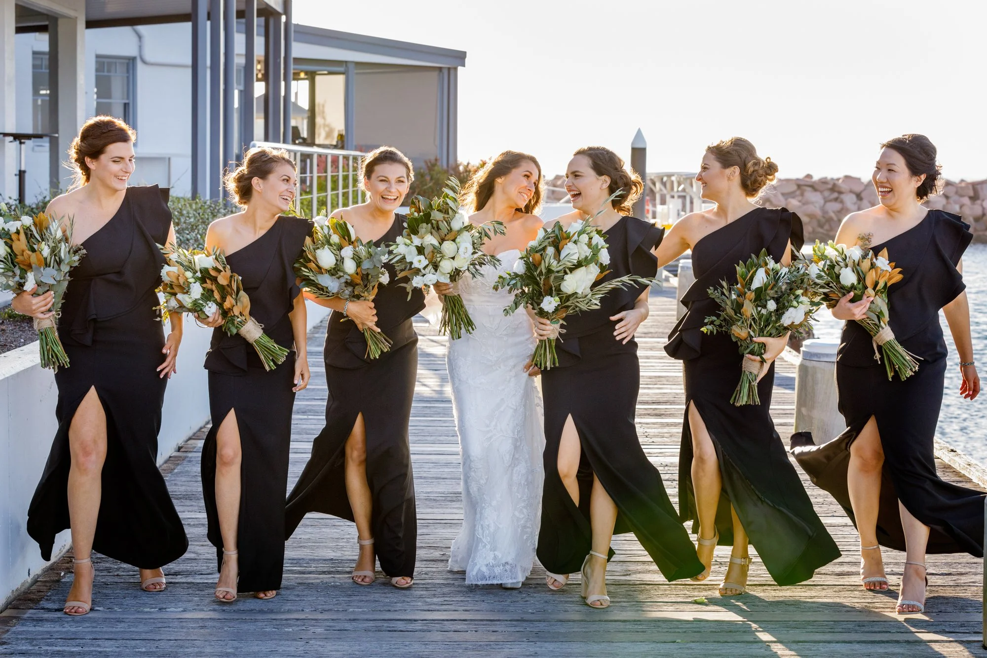 A bride and seven bridesmaids walking on a wooden dock by the water, holding bouquets of white and green flowers, all smiling and enjoying each other's company in the late afternoon sunlight.