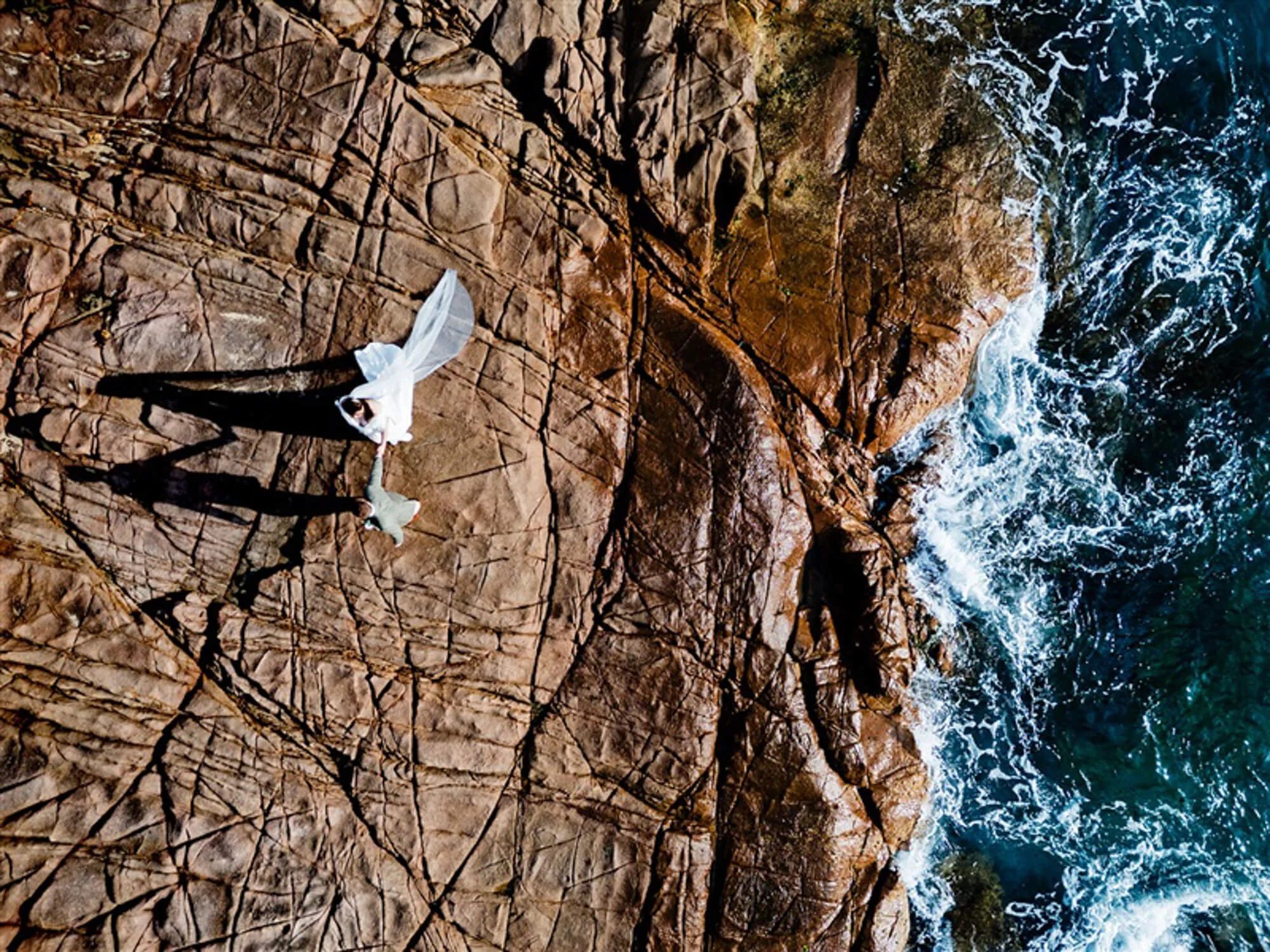 A couple dressed in white standing on rocky shoreline with ocean waves crashing nearby, casting long shadows on the rocks.