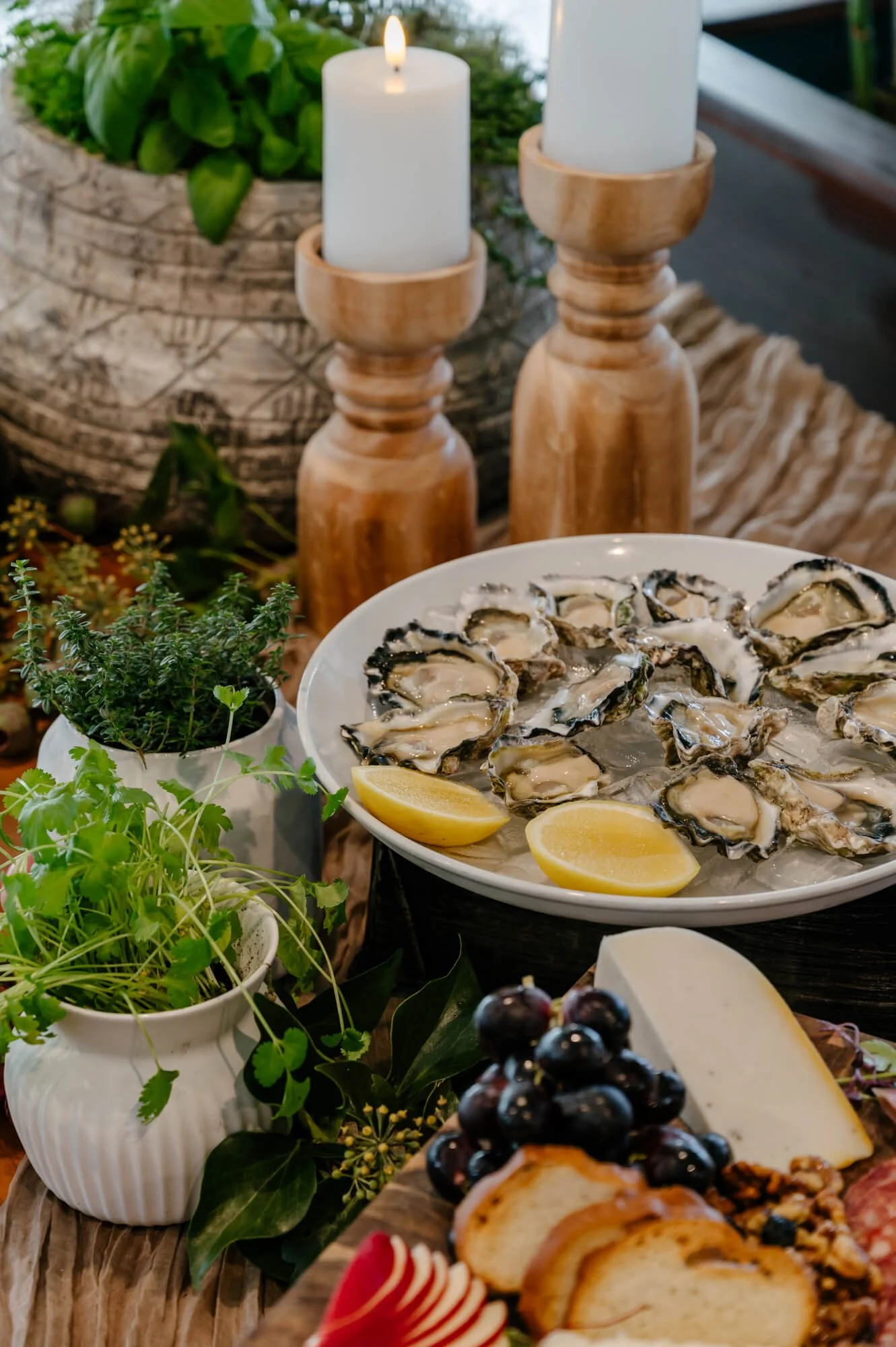 Plate of fresh oysters with lemon wedges on ice, surrounded by herbs, candles, and a cheese platter.