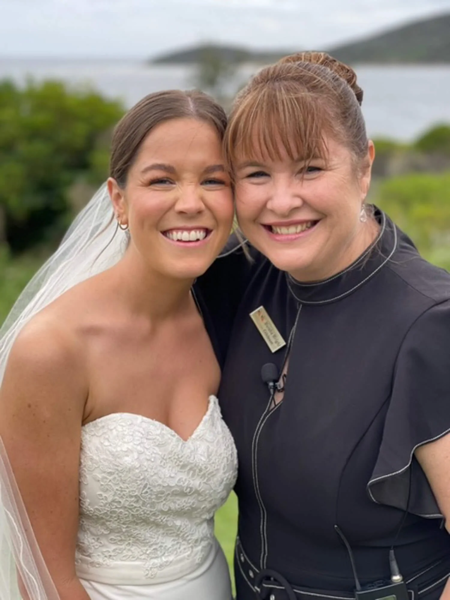 Two women smiling outdoors, one in a wedding dress and the other in a black top, with a scenic background of green trees and water.