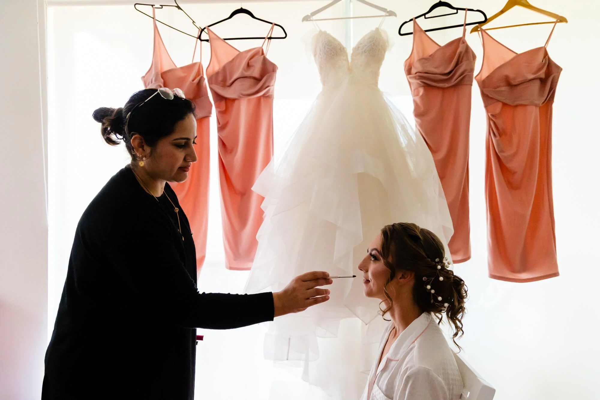 A woman is applying makeup to another woman, who is seated. Several peach-colored dresses are hanging on hangers in the background, along with a white wedding gown.