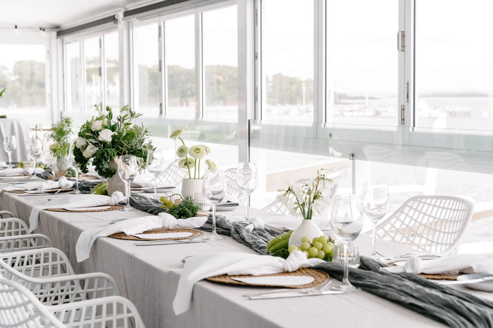 Elegant table set for a meal with white tablecloth, white cloth napkins, green and white floral centerpiece, glassware, and woven placemats, in a bright room with large windows overlooking a marina.