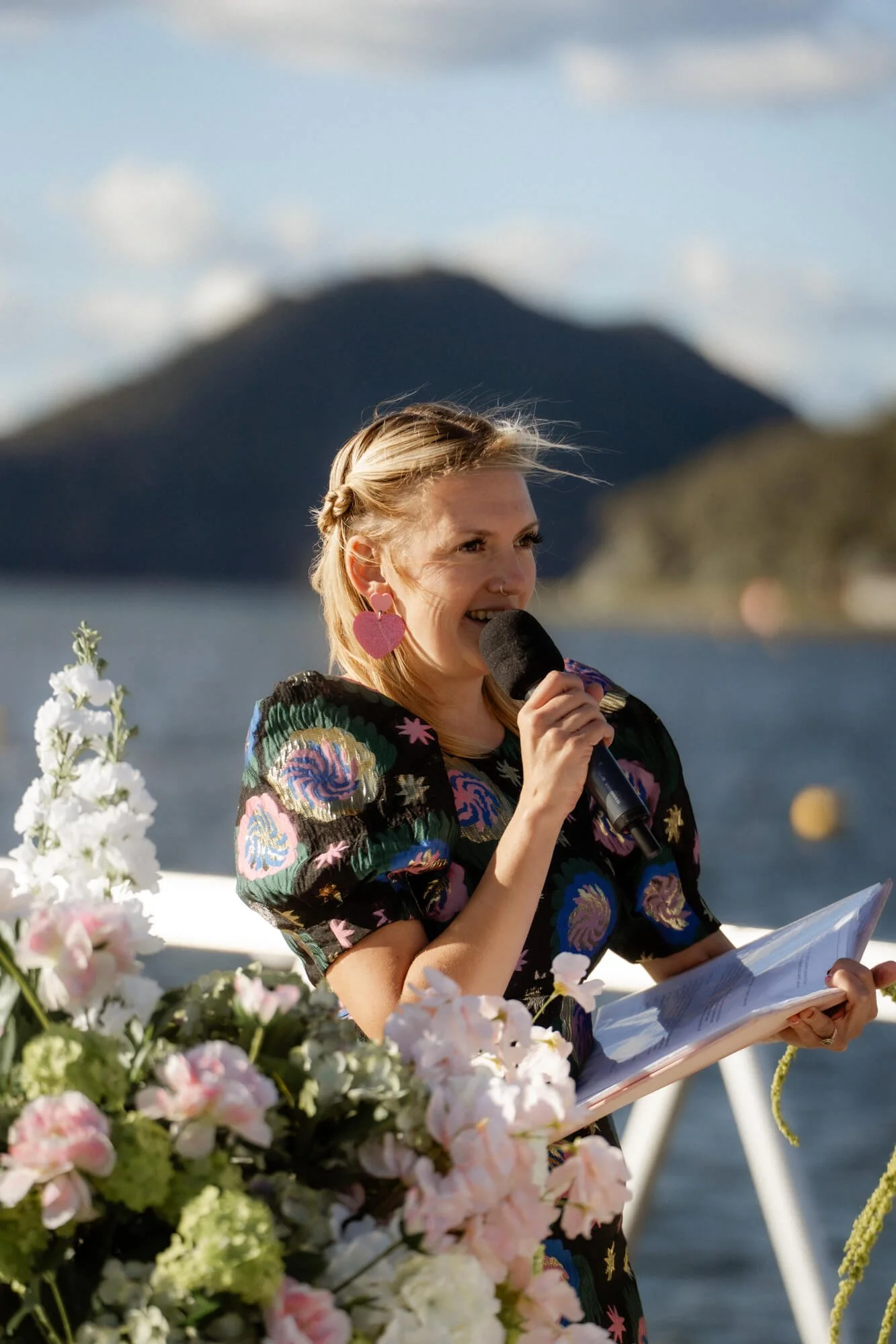 A woman with blonde hair in a braid, wearing large pink heart-shaped earrings and a colorful patterned dress, holding a microphone and reading from a paper, standing near a body of water with mountains in the background, decorated with flowers.