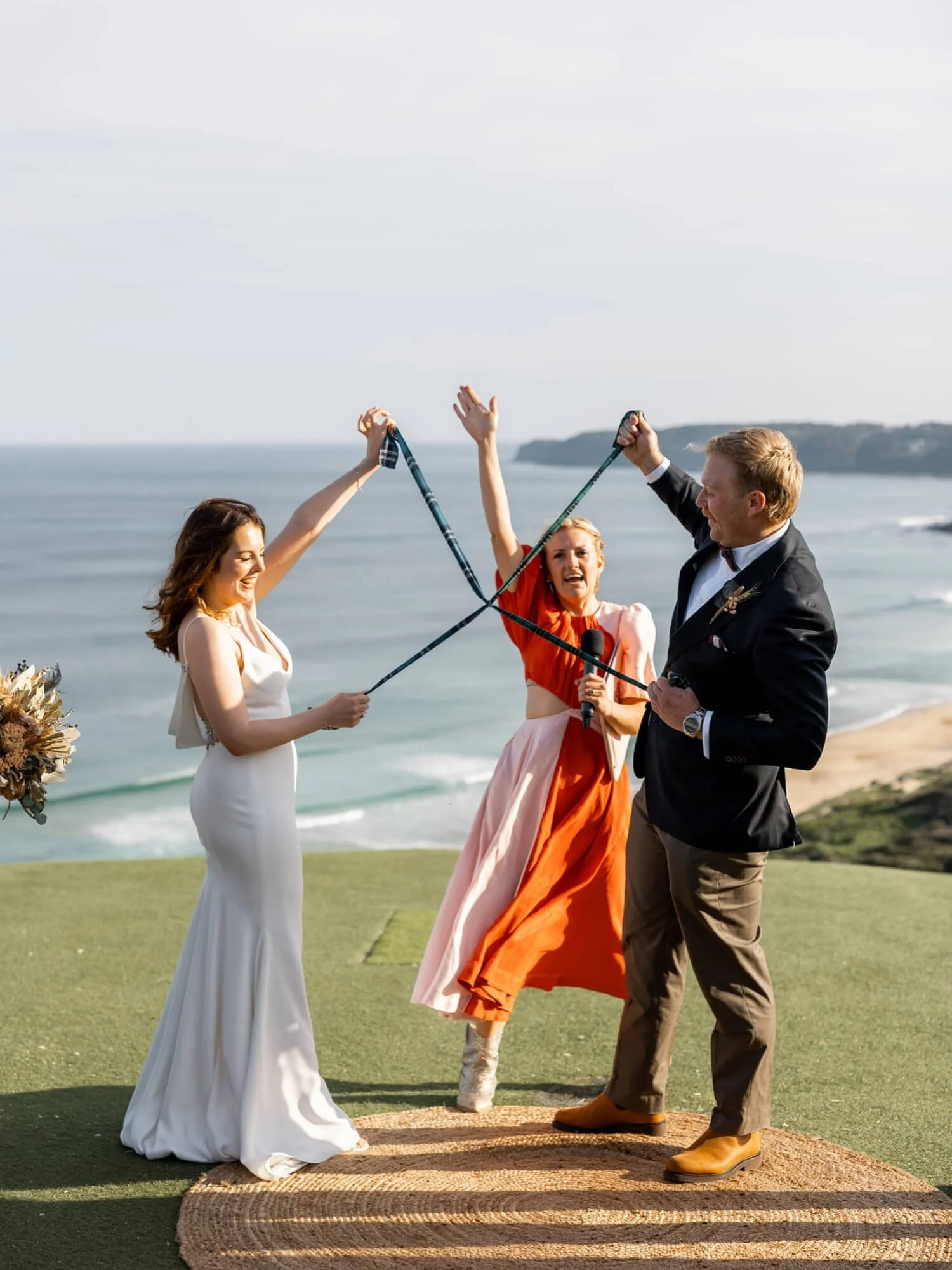 A wedding ceremony on a grassy cliff overlooking the ocean. The bride in a white dress and the groom in a black jacket and tan pants are celebrating with a woman in an orange dress. The bride and woman are holding ribbons, and the woman is holding a 