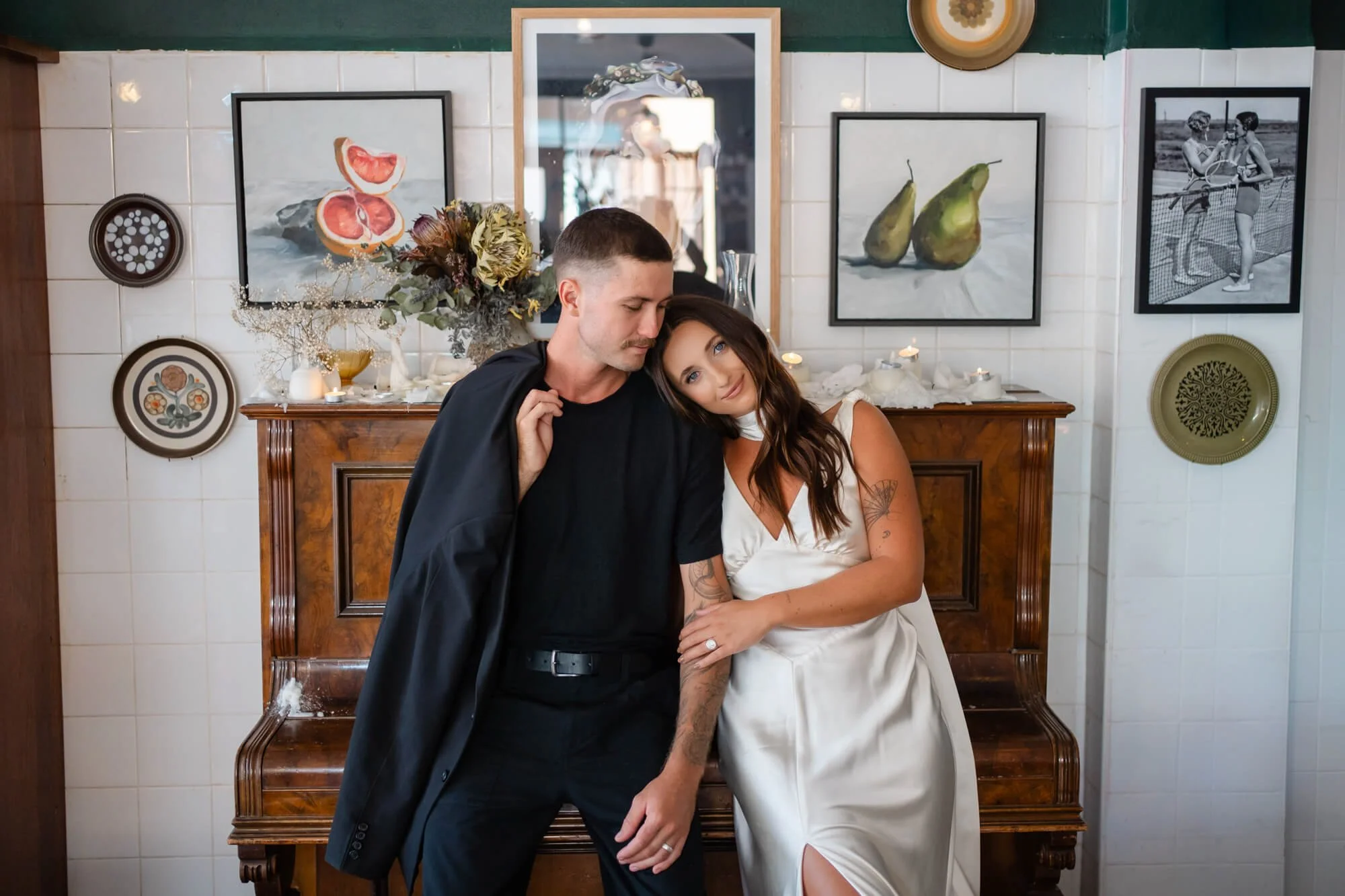 A couple stands close together, the woman leaning her head on the man's shoulder, in front of a wooden cabinet with decorative plates, candles, and a bouquet of flowers. The background features framed artwork and a mirror on a white tiled wall.