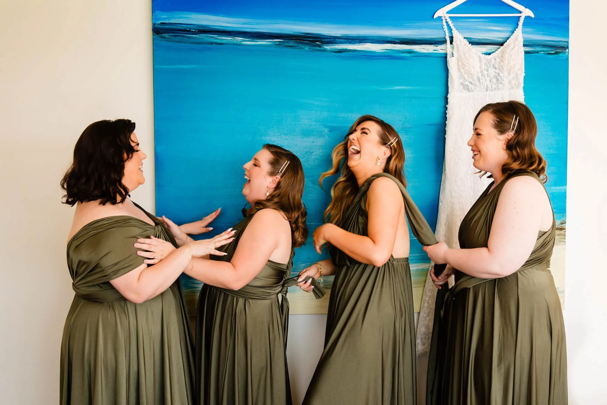 Four women in matching olive green dresses laughing and smiling together, indoors with a beach scene painting and a white dress hanging in the background.