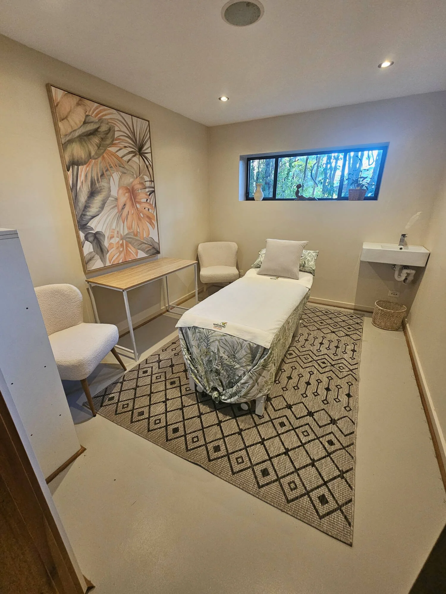 Small room with a single bed, two chairs, a desk, a wall painting, a window with decor, and a small sink in the corner.