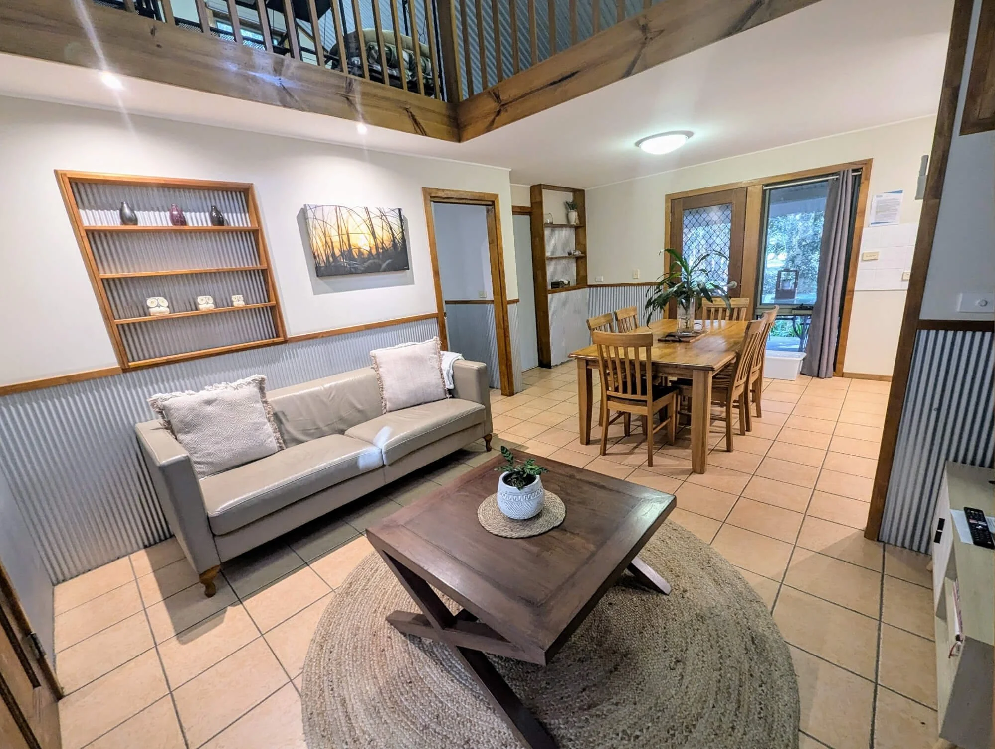 Living and dining area with a beige couch, wooden coffee table, and a wooden dining table with six chairs. There is a potted plant on the table, and the space features wooden accents, shelves, and a sliding glass door leading outside.