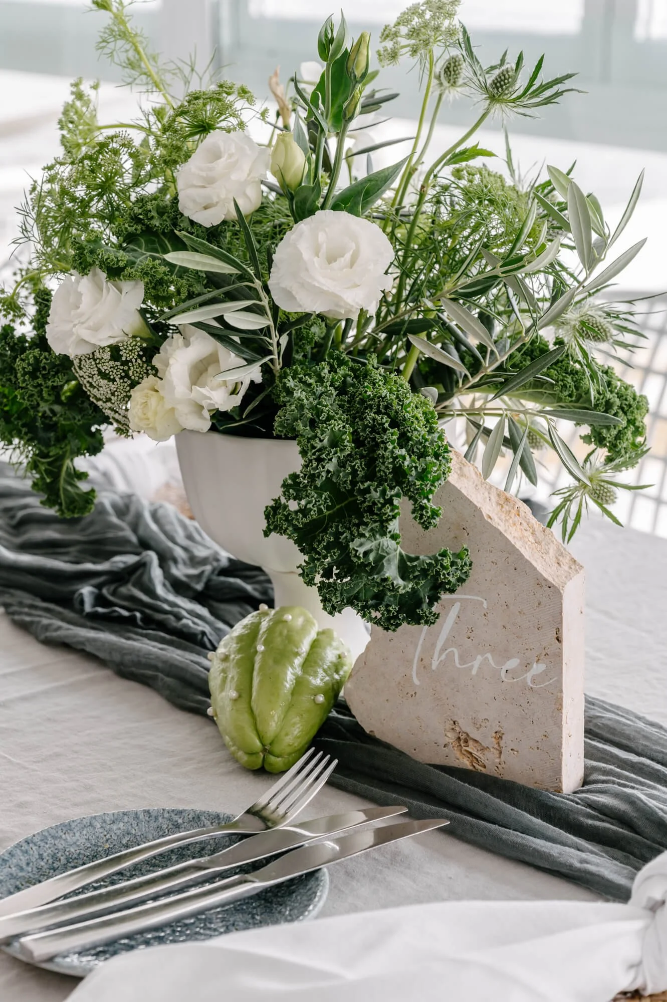 A floral centerpiece with white flowers and greenery in a white vase on a table, with a small decorative stone piece reading 'three', a green ornamental gourd, and place settings including a blue plate and silverware, on a table covered with a light-
