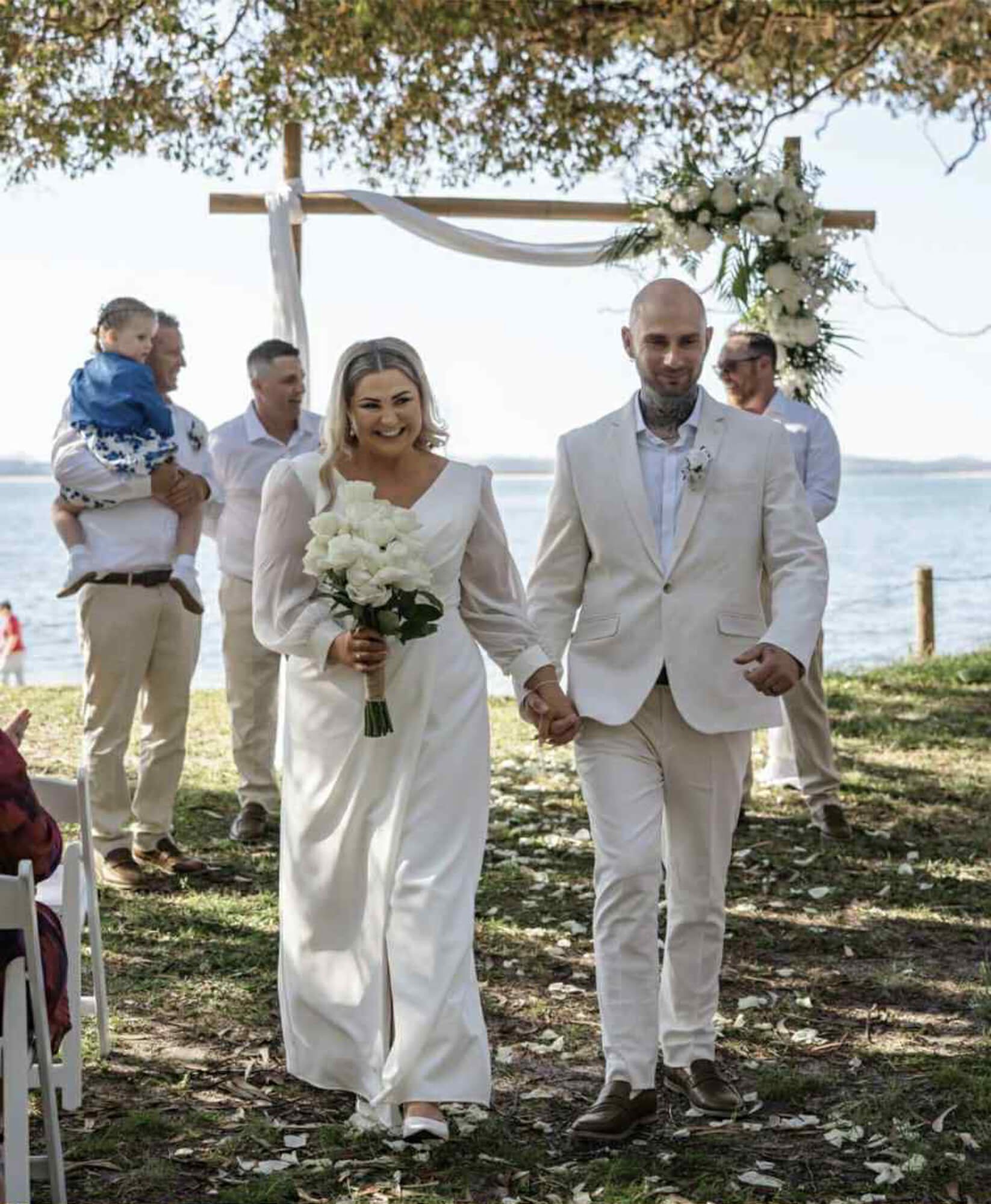 A newlywed couple walks hand-in-hand outdoors near water, smiling, with the bride holding a bouquet of white flowers and the groom wearing a white suit. In the background, a small group of people is gathered, and a wedding arch with white drapery and