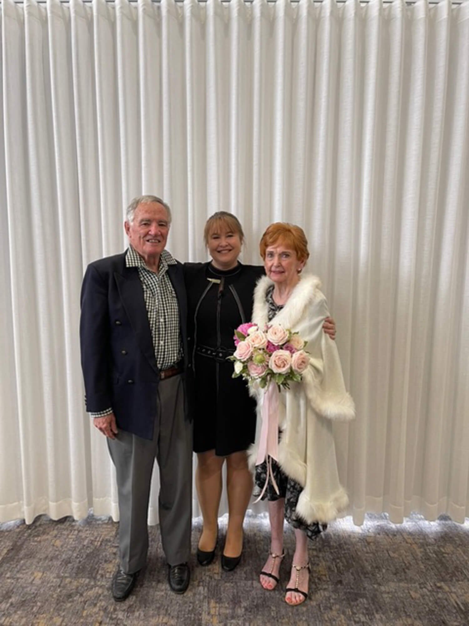 Three people smiling in front of a white curtain, one holding a bouquet of pink and white roses, dressed in formal and semi-formal attire.