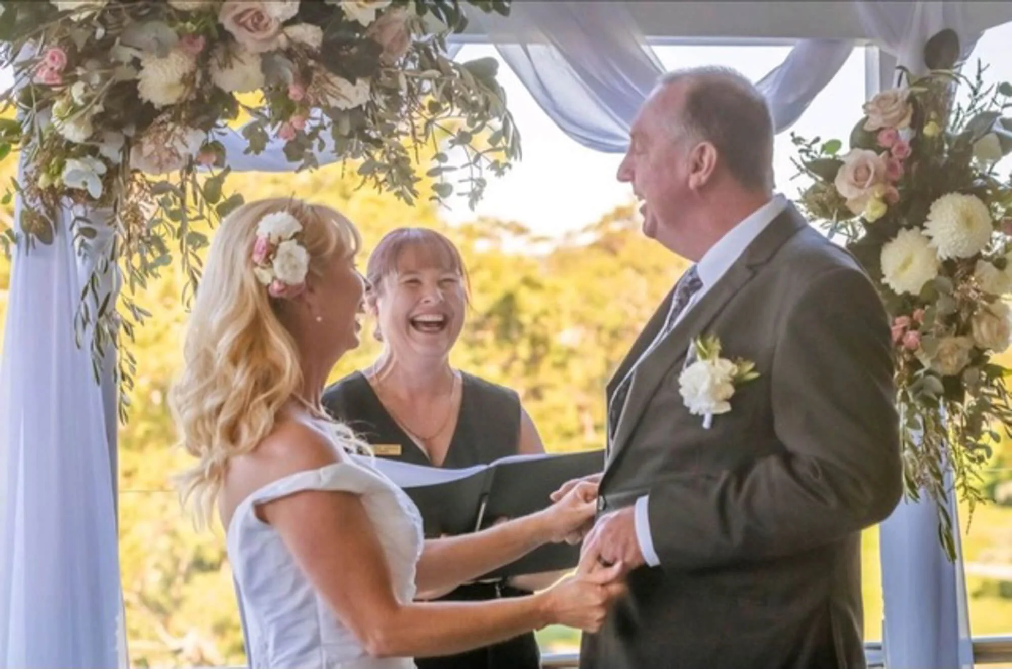 A bride and groom holding hands and smiling at each other during a wedding ceremony under a floral arch, with an officiant standing behind them, outside in a sunny setting.