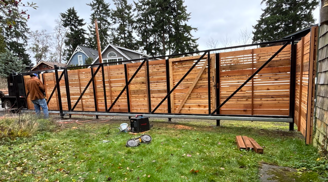 A person working on a wooden fence with metal framework, using power tools in a backyard with grass and trees, and houses in the background.