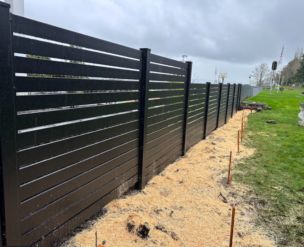A black horizontal slat fence along a grassy area with some construction stakes and supplies nearby. The sky is cloudy.