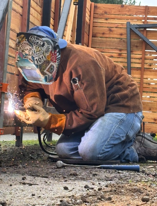 A person wearing a welding helmet, brown jacket, jeans, and gloves kneels on the ground while welding a metal piece in an outdoor setting. Sparks are visible from the welding process.