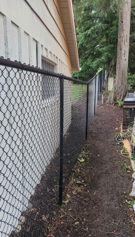 A black chain-link fence running alongside a beige house with a window, with trees and a dirt pathway on the other side.