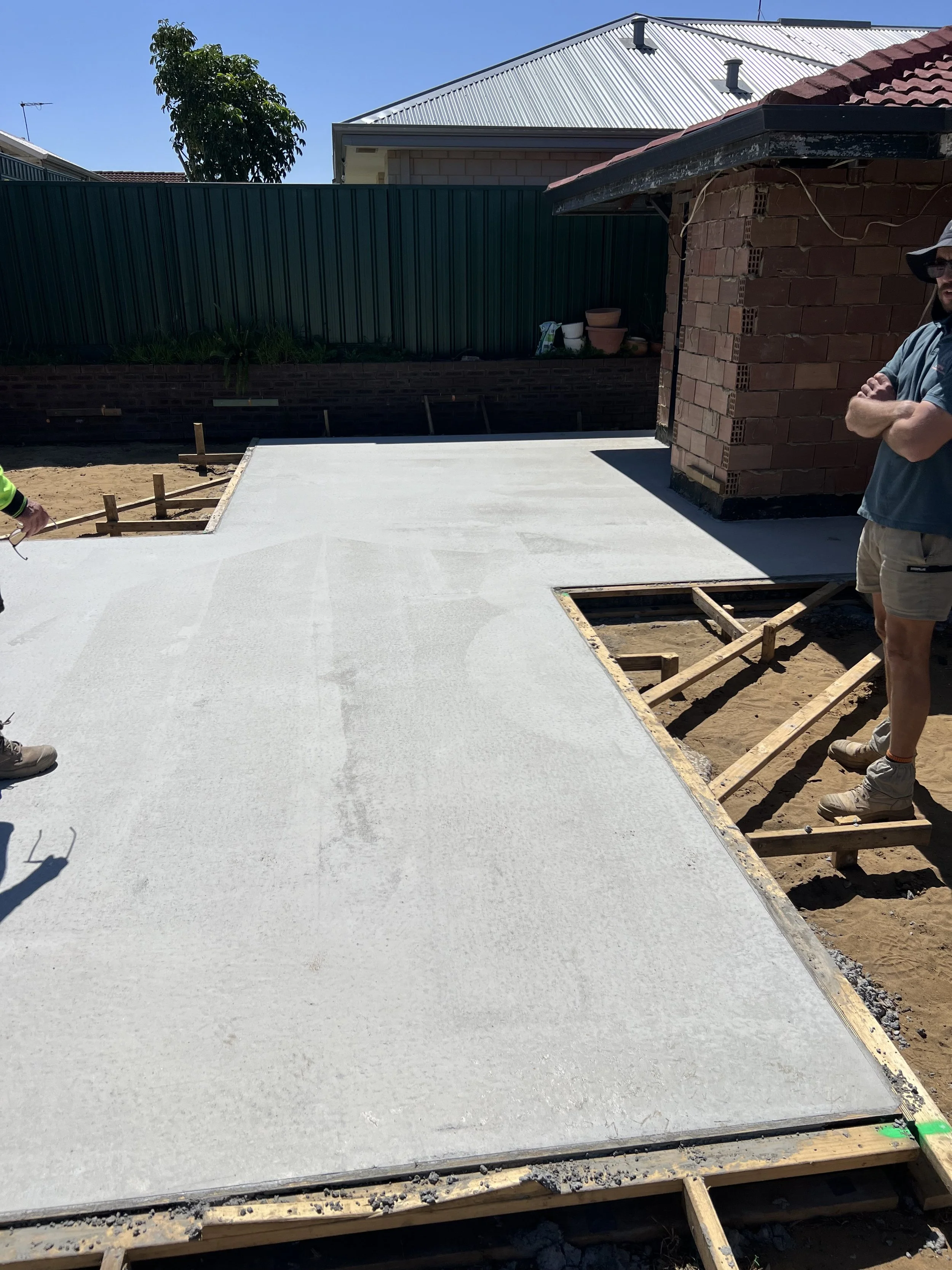 Construction site with a partially finished concrete slab, next to an unfinished brick structure, and a man standing nearby wearing a gray shirt and shorts, on a sunny day.