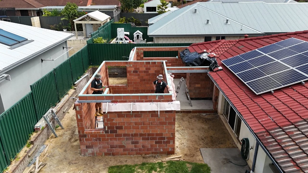 Construction site with a brick structure being built next to a house with solar panels on its red roof. Two workers are on the brick walls, one holding tools and the other standing on a ladder.