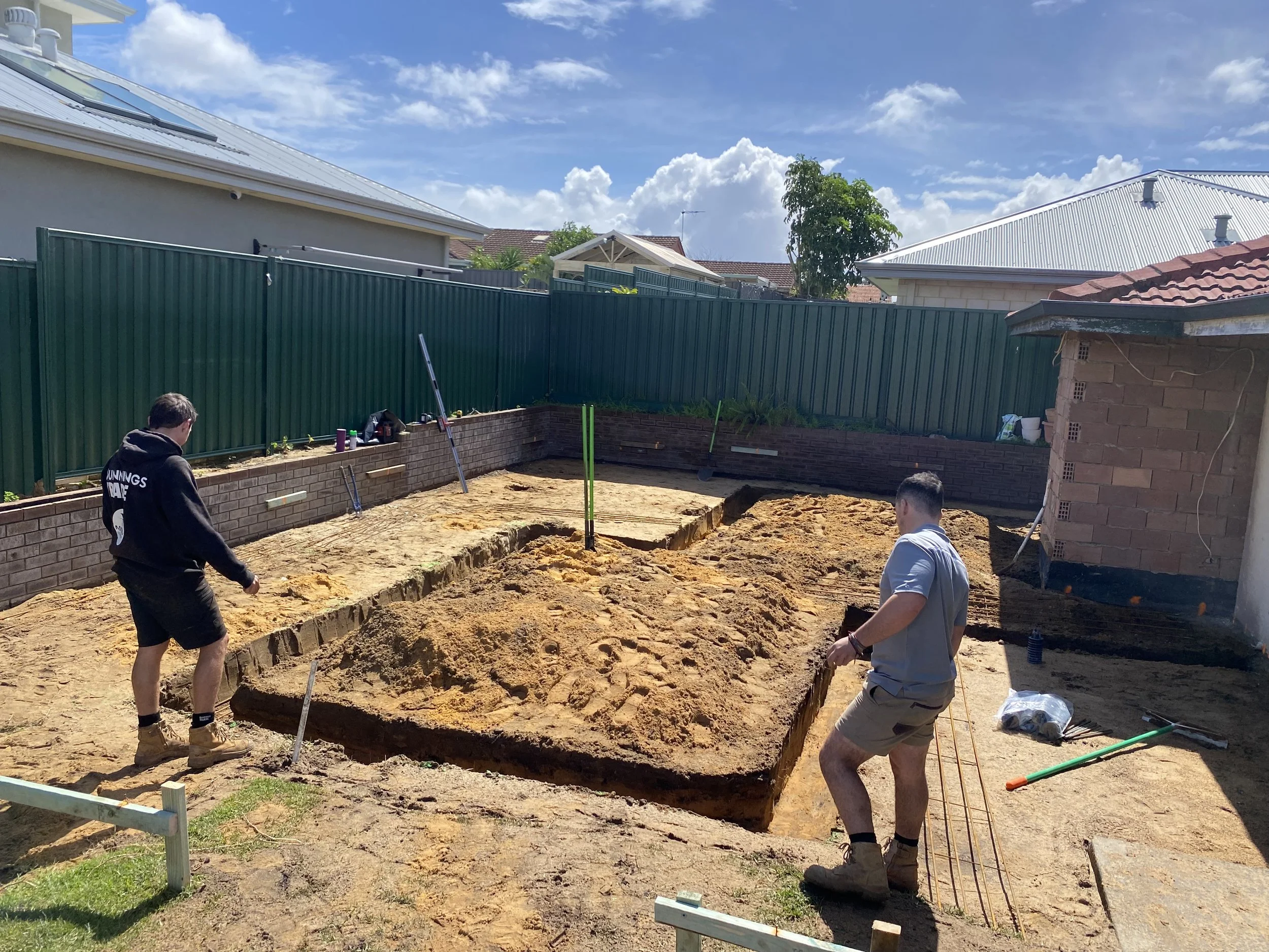 Construction site with two workers digging a trench in the backyard of a residential area under a partly cloudy sky.