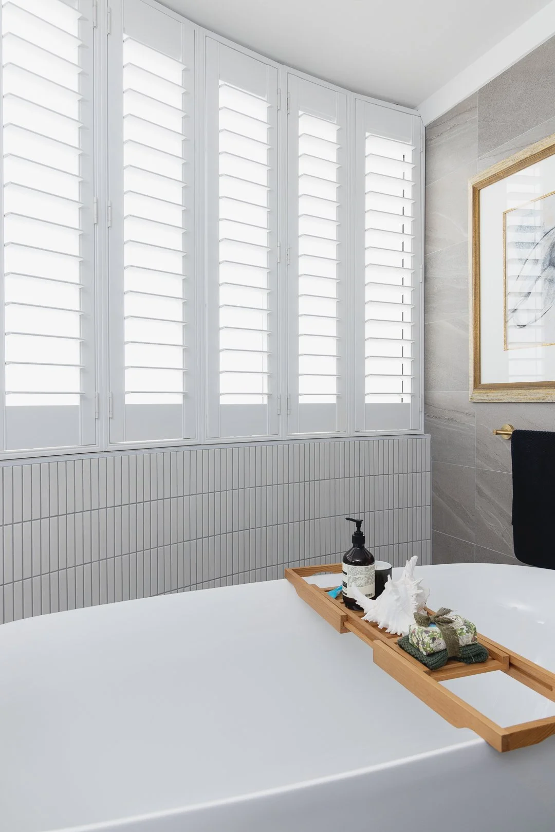Bathroom with white shuttered windows, gray tiled wall, and a white bathtub with a wooden tray holding toiletries and towels.