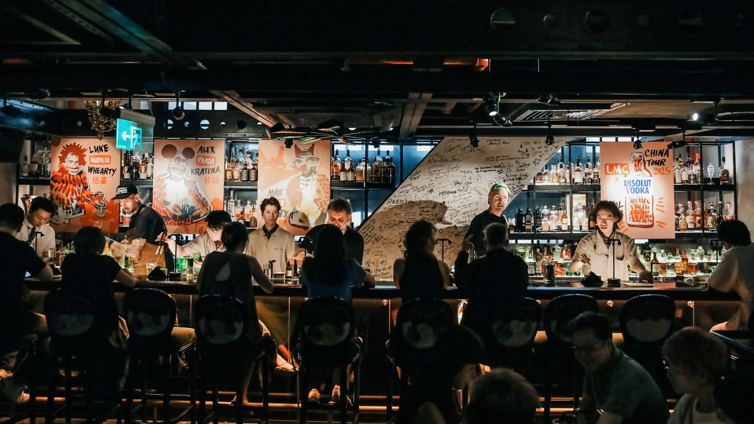 People sitting at Obsidian bar in Shenzhen, China during the LMC Vol #23 event. Four orange posters are on display behind the bar with collage-style images of Luke Whearty, Alex Kratena, Matt Whiley and Absolut Vodka bottle.
