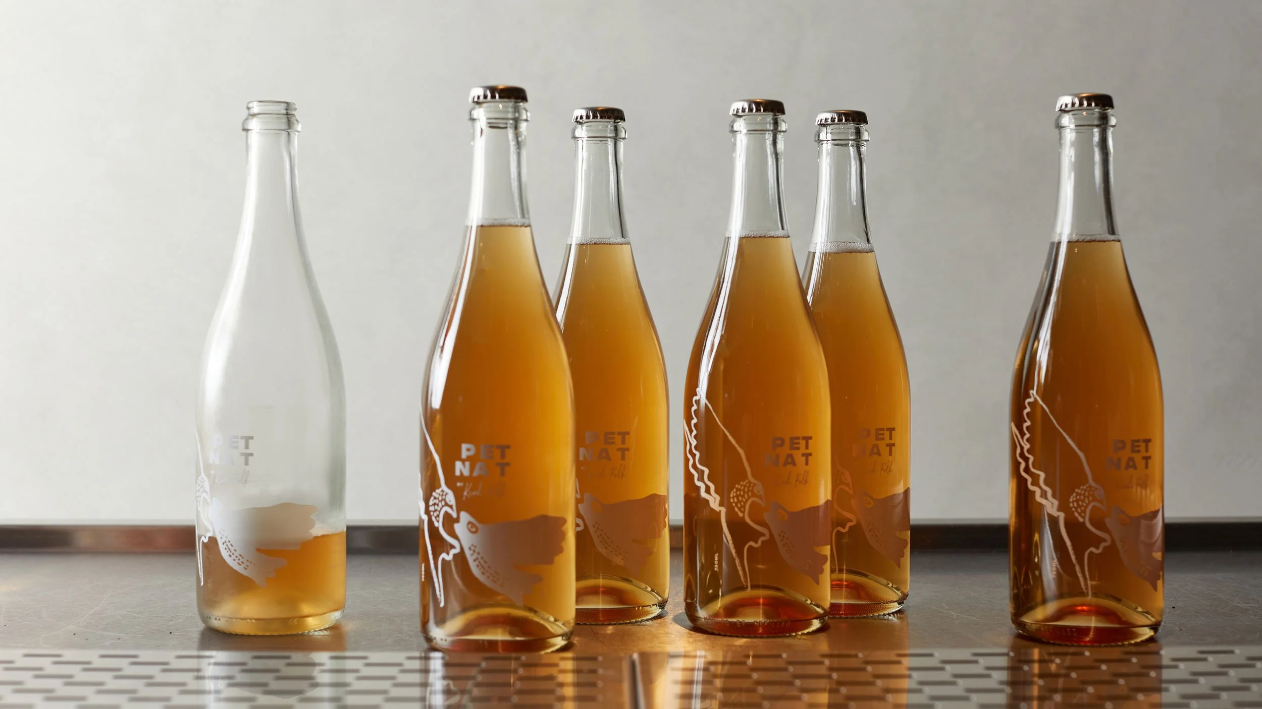 Six bottles of Byrdi x Kind Folk Pet Nat sitting on a grey surface. Left hand bottle is open and half empty.