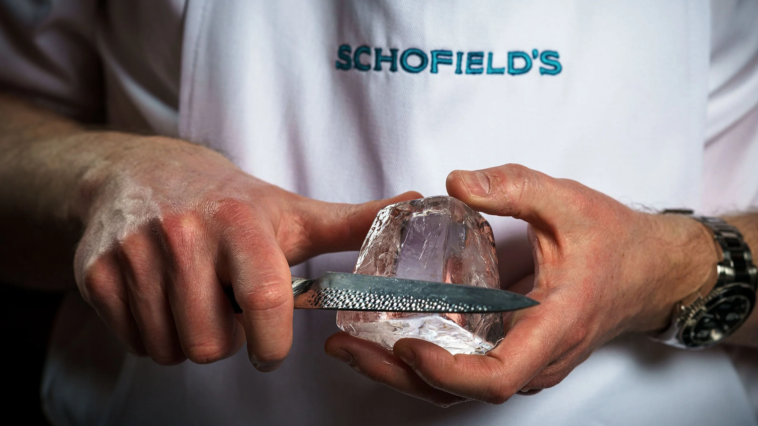 Hands of Joe Schofield use a knife to carve a block of ice. Schofield’s teal logo can be seen embroidered on his white apron.