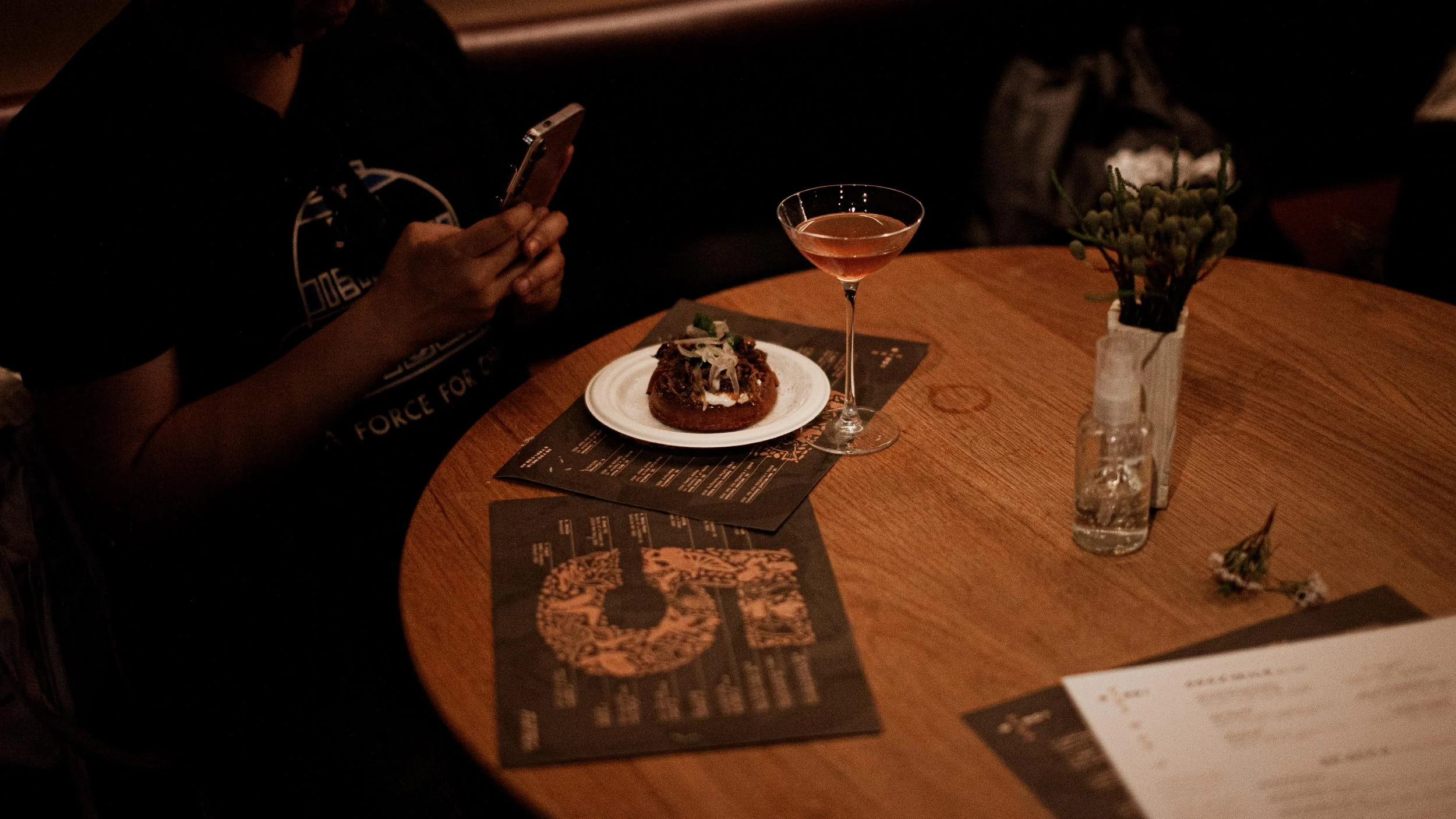 A person sitting at a wooden table looking at their phone. Menus, a snack and a cocktail in a martini glass are on the table.