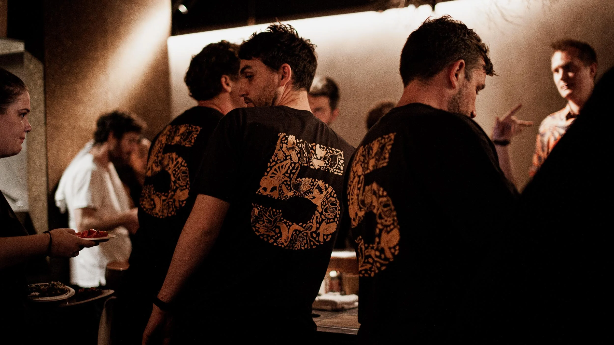 Rear view of three people standing at the Byrdi bar, they are all wearing black tshirts with Illustrated birds in a 5 shape on the back in mustard.