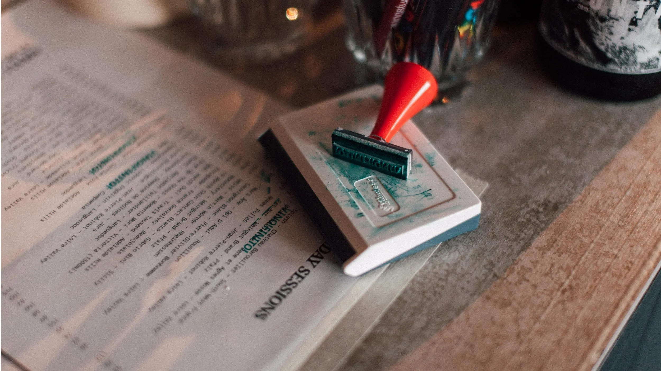 Close-up of a stamp resting on a stamp pad with the words “Vino Finito” visible. The stamp pad sits on a Someday Sessions wine menu with some of the wines stamped out in green.