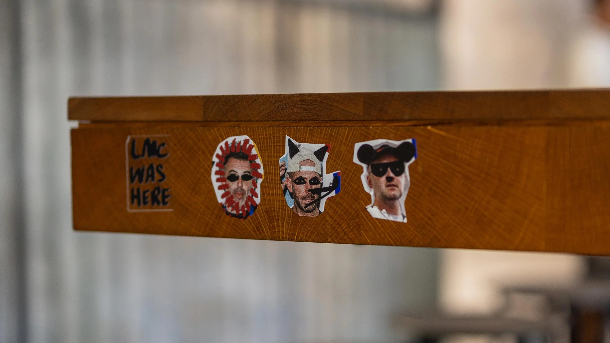 Four stickers pasted to the side of a wooden table. “LMC WAS HERE” and heads of Luke Whearty, Matt Whiley and Alex Kratena.