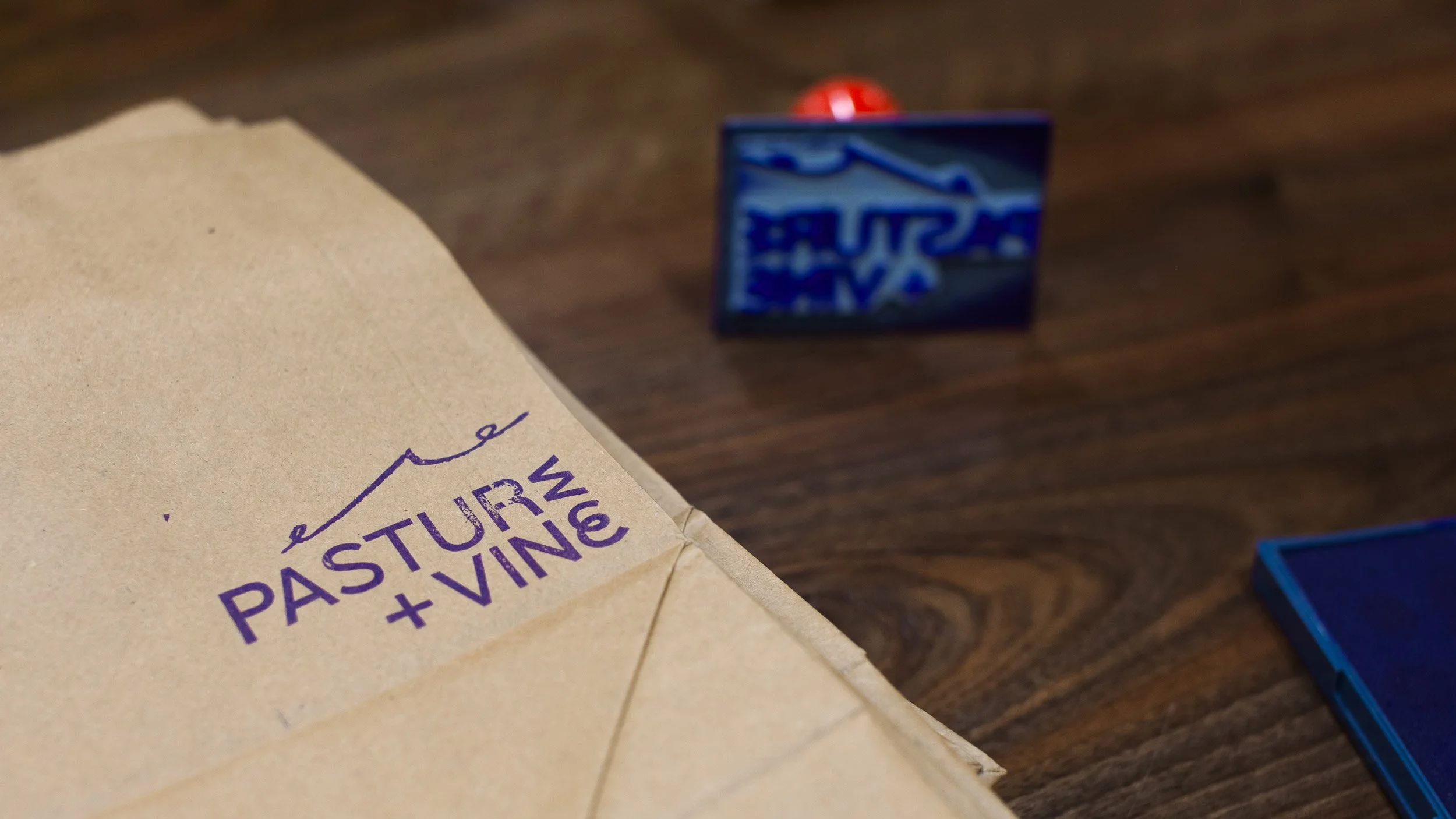 Closeup up Pasture and Vine logo stamped onto a brown paper bag in purple ink. Rubber stamp sits in background.