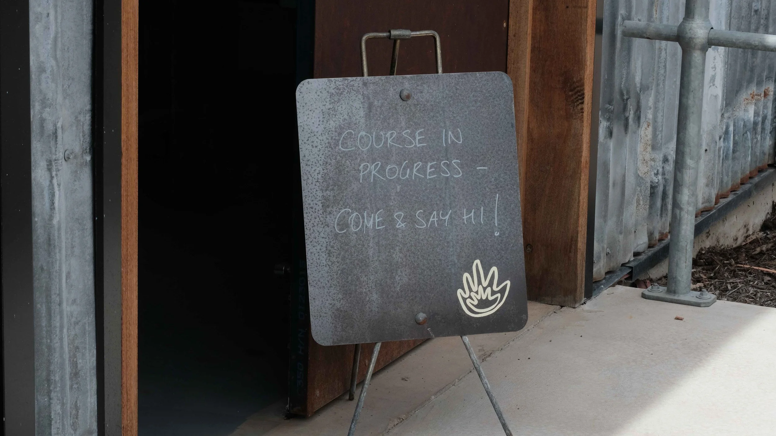 Patinaed metal rectangular A-Frame sitting on concrete in front of an open door reading “Course in Progress – Come and Say Hi!”. In the bottom right hand corner of the sign is the Two Hands Forge logo in pale yellow.
