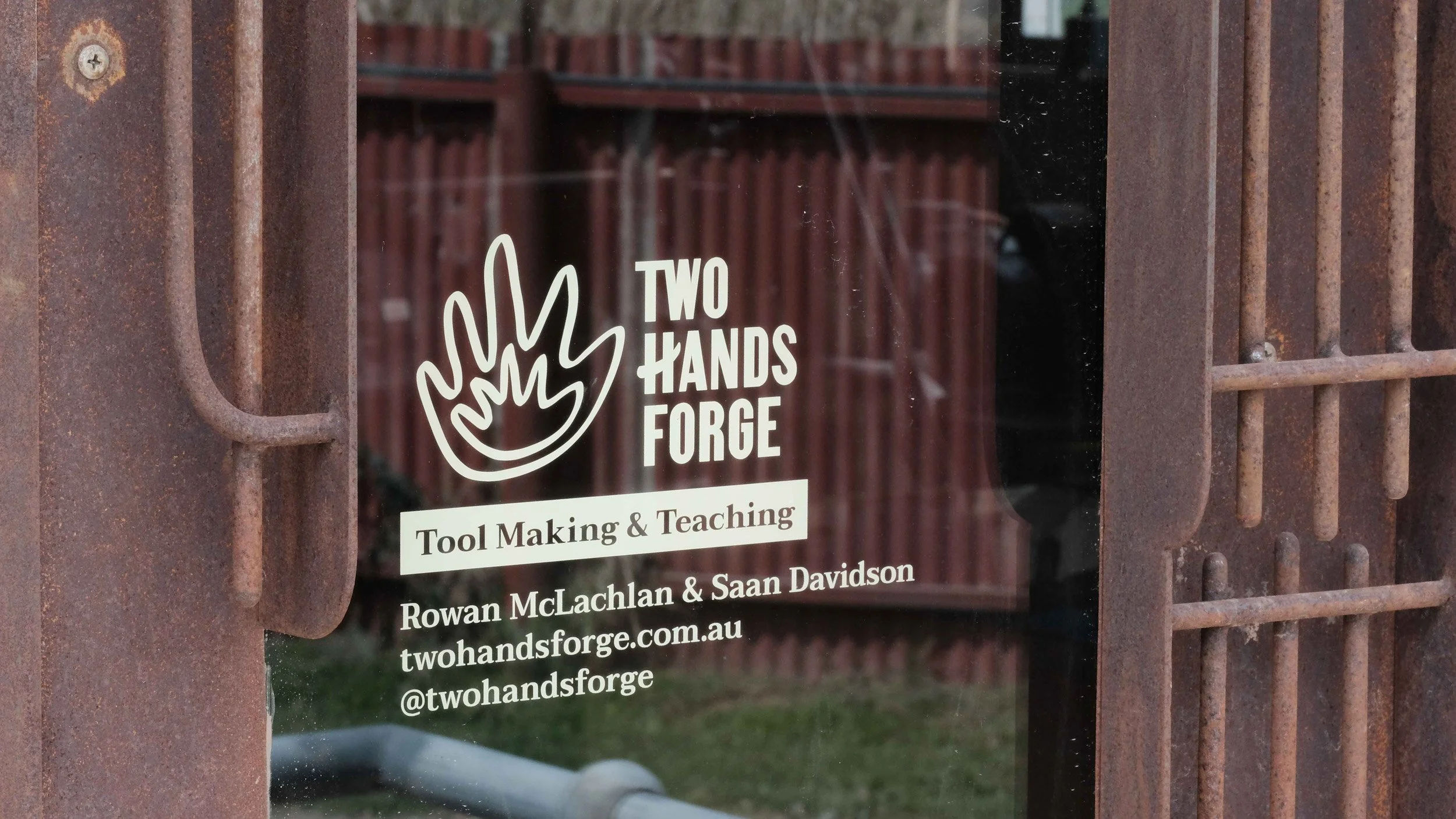 Closeup of decal on glass door featuring Two Hands Forge logo, “Tool Making & Teaching” and the website and instagram handle.