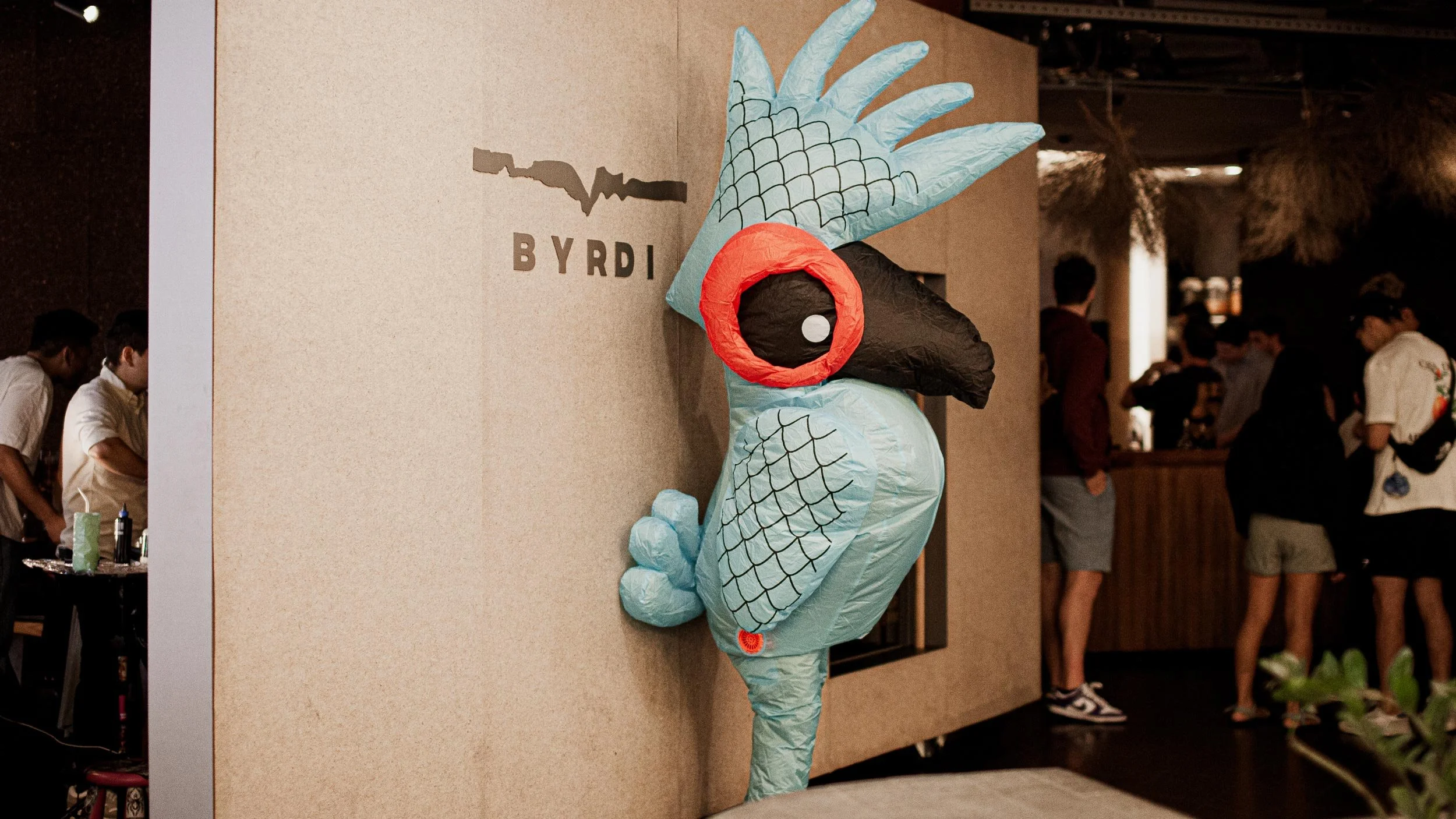 A person in an inflatable blue parrot costume stands in front of the entrance of Byrdi bar. We can people inside Byrdi through the door.