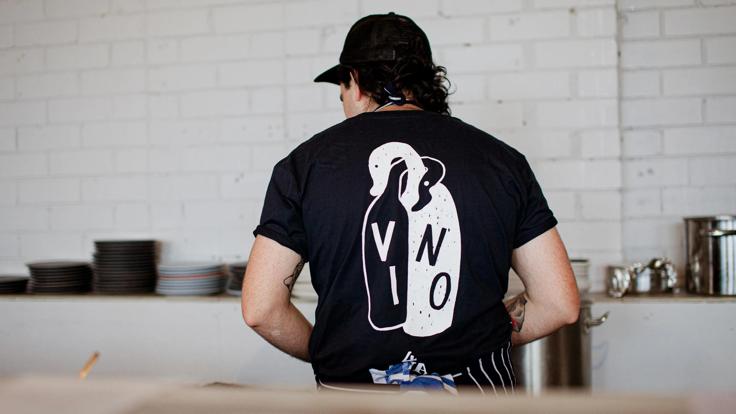 Rear view of Pete Cooksley wearing navy Someday Sessions t-shirt with “Vino Forever” illustration in white features.