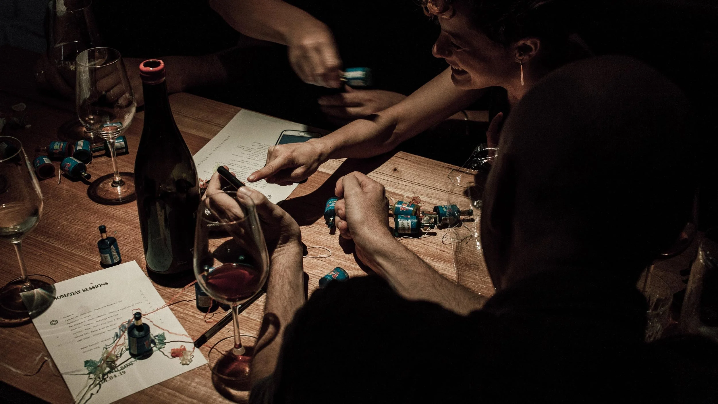People gathered around a wooden table pointing at something on a mobile phone at Someday Sessions at Osteria Ilaria in Melbourne. Party poppers, wine glasses and Someday Sessions menus are on the table.