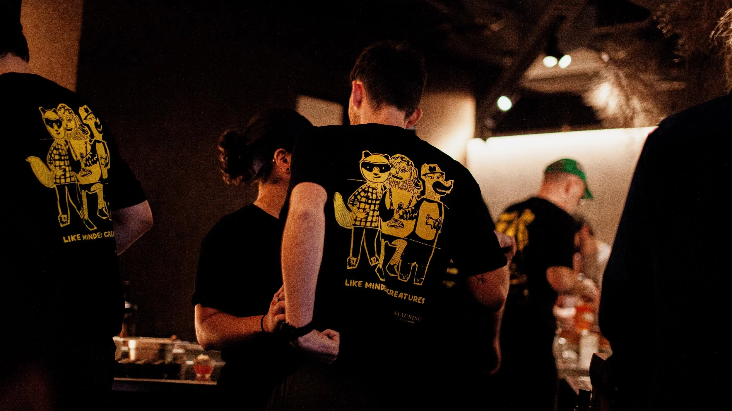 Rear view of people standing at the Byrdi bar, they are all wearing black t-shirts with an exquisite corpse illustration made up of a panda, a fox and a wookie on the back in yellow and the words “Like Minded Creatures”.