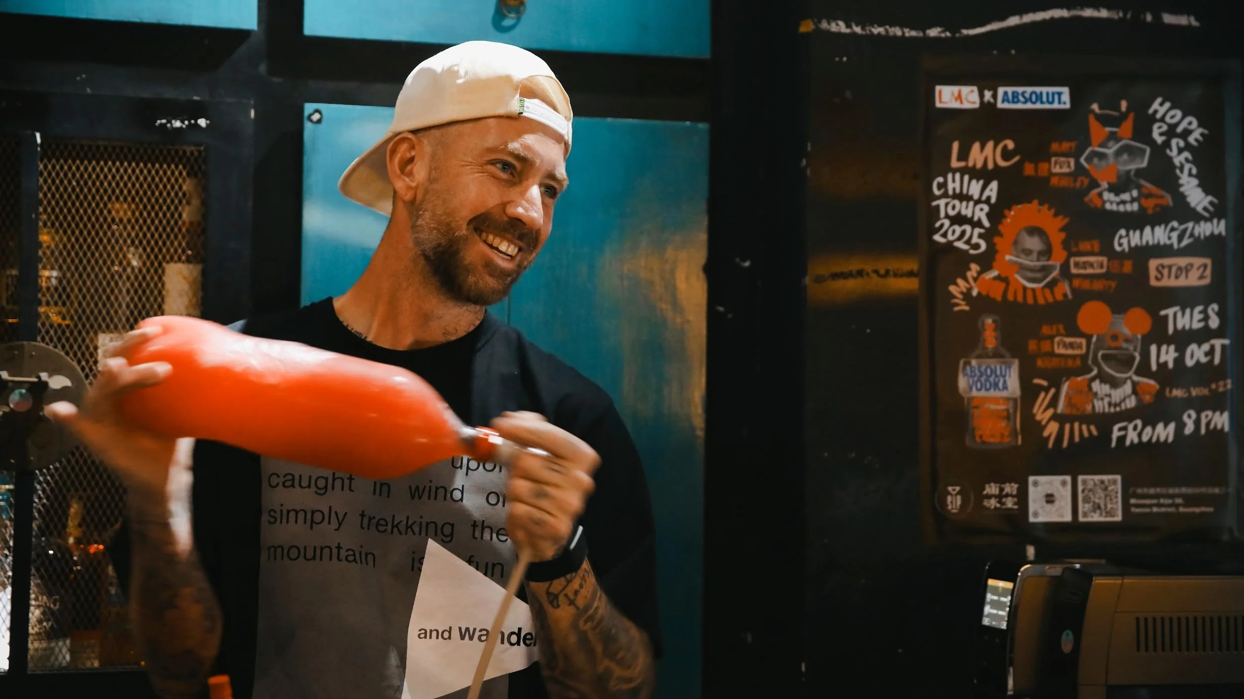 Matt Whiley smiling in a white cap holding a bottle of orange liquid. In the background is a black poster with collage illustrations and hand drawn type promoting the LMC Vol #22 event.