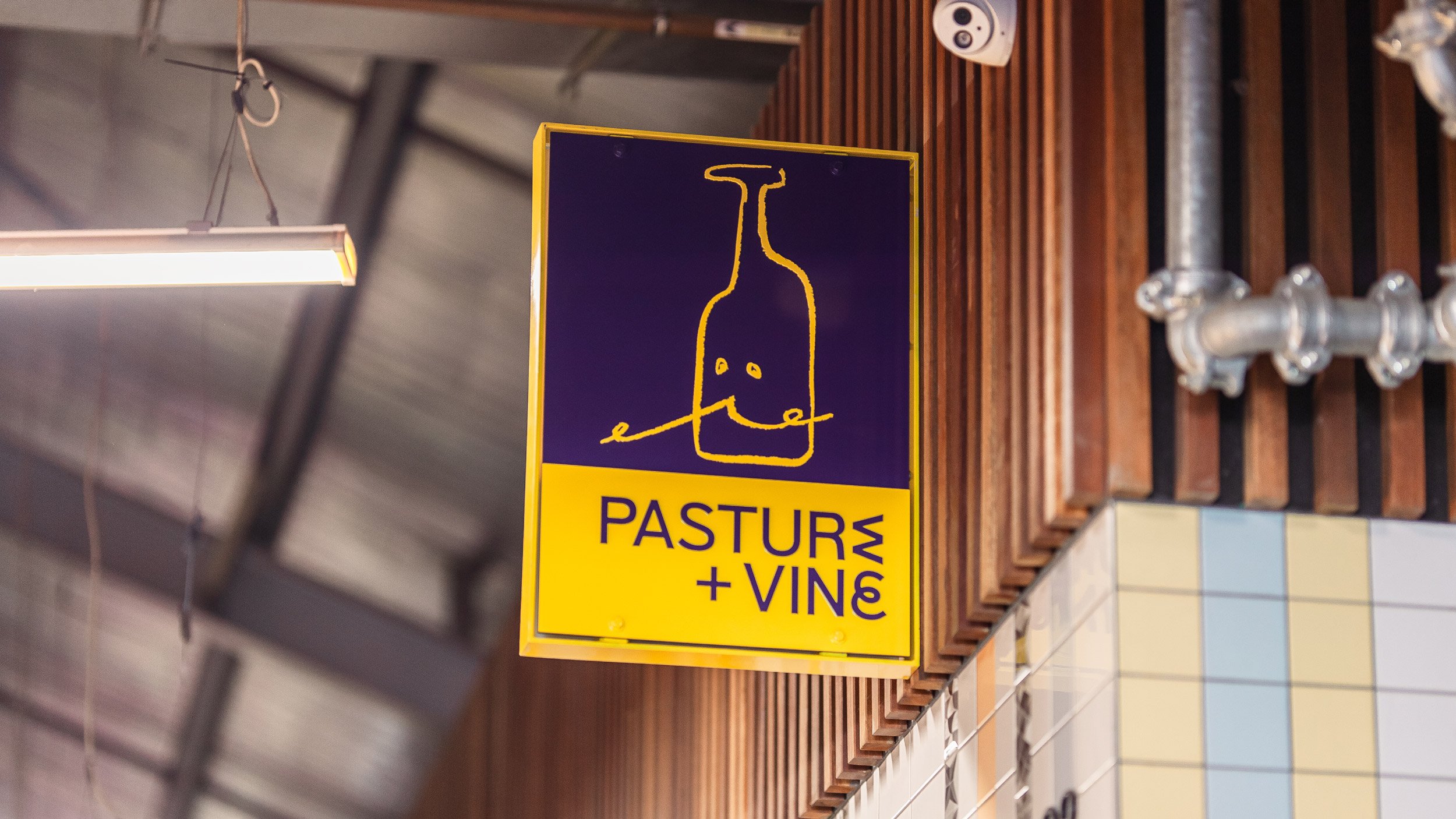 Closeup of Pasture and Vine acrylic signage showing illustration of wine bottle caricature with moustache in yellow on purple with logotype below in purple on yellow.