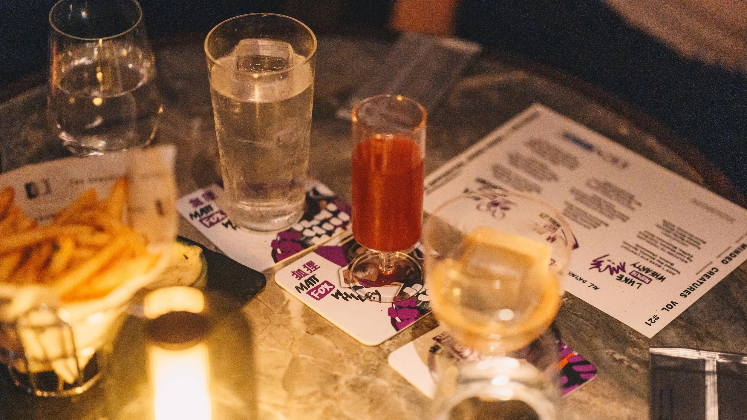 Three cocktails sit on top of LMC coasters on a grey marble table alongside a menu for Like Minded Creatures Vol #21.