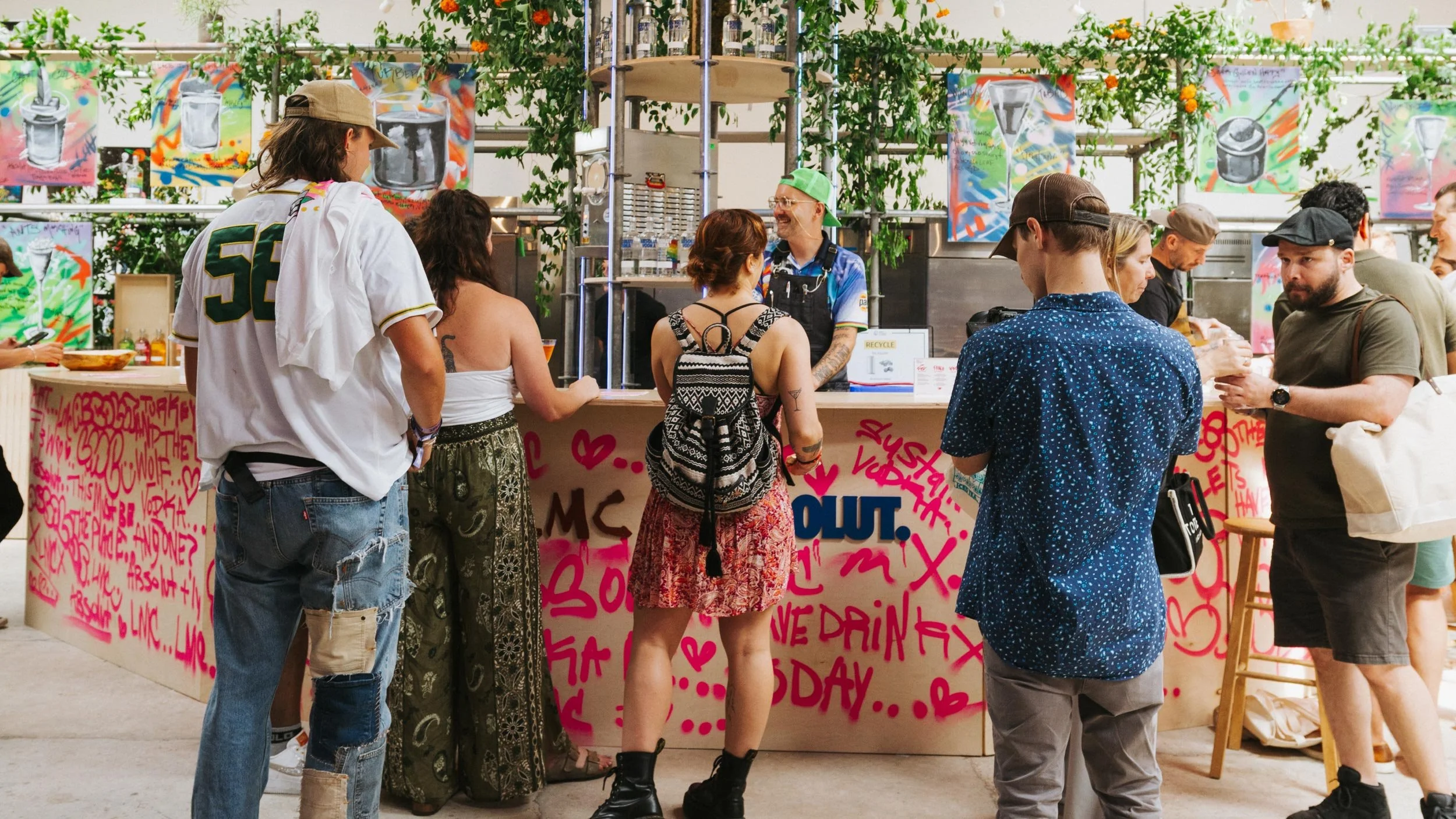 People at a wooden bar with LMC x Absolut logo lockup on the front. Pink spray painted words cover the bar. Bartenders Alex Kratena and Matt Whiley are preparing drinks.