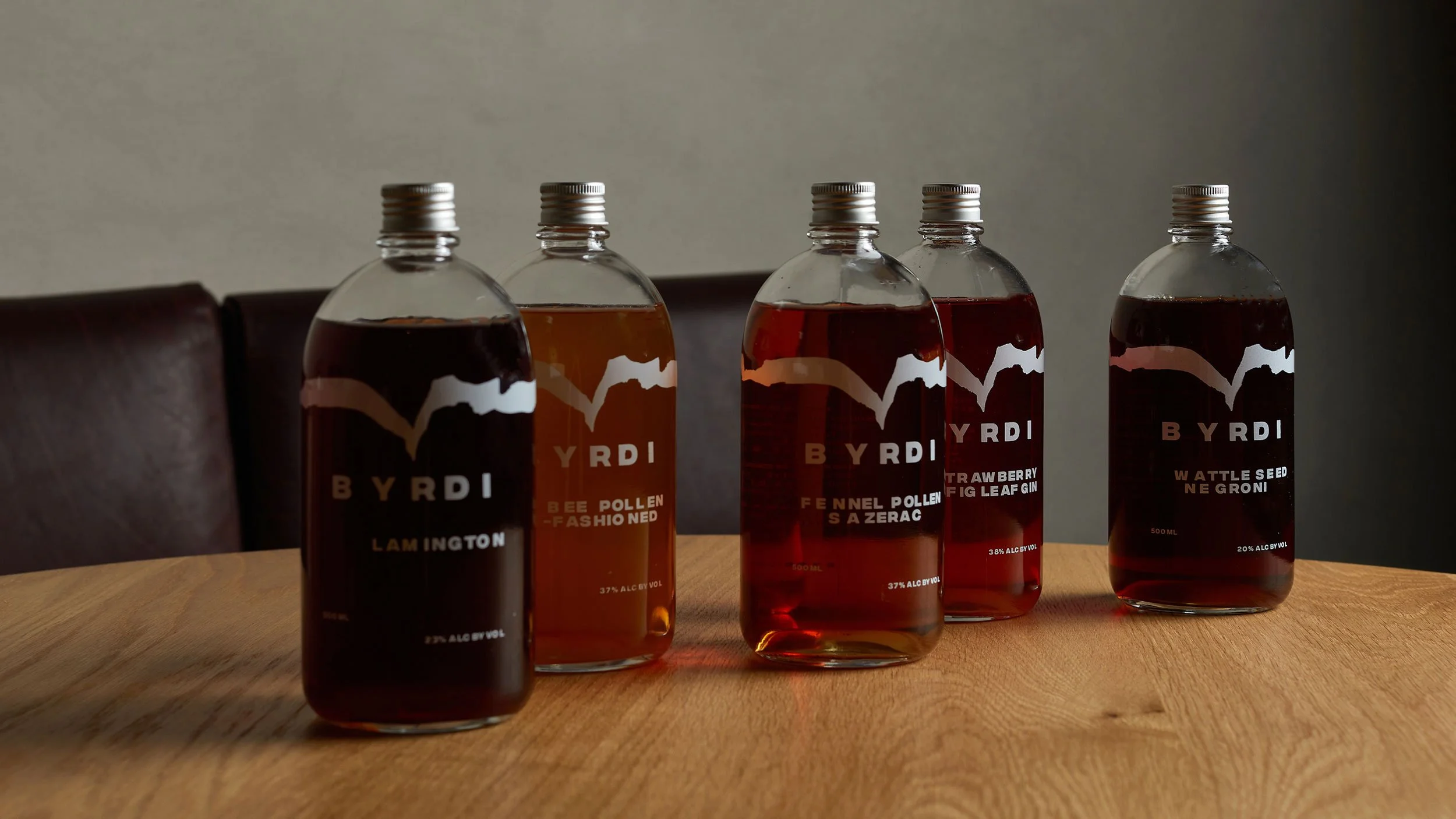 Five Byrdi bottled cocktails sitting on a wooden table. The cocktails are in shades of orange and red.