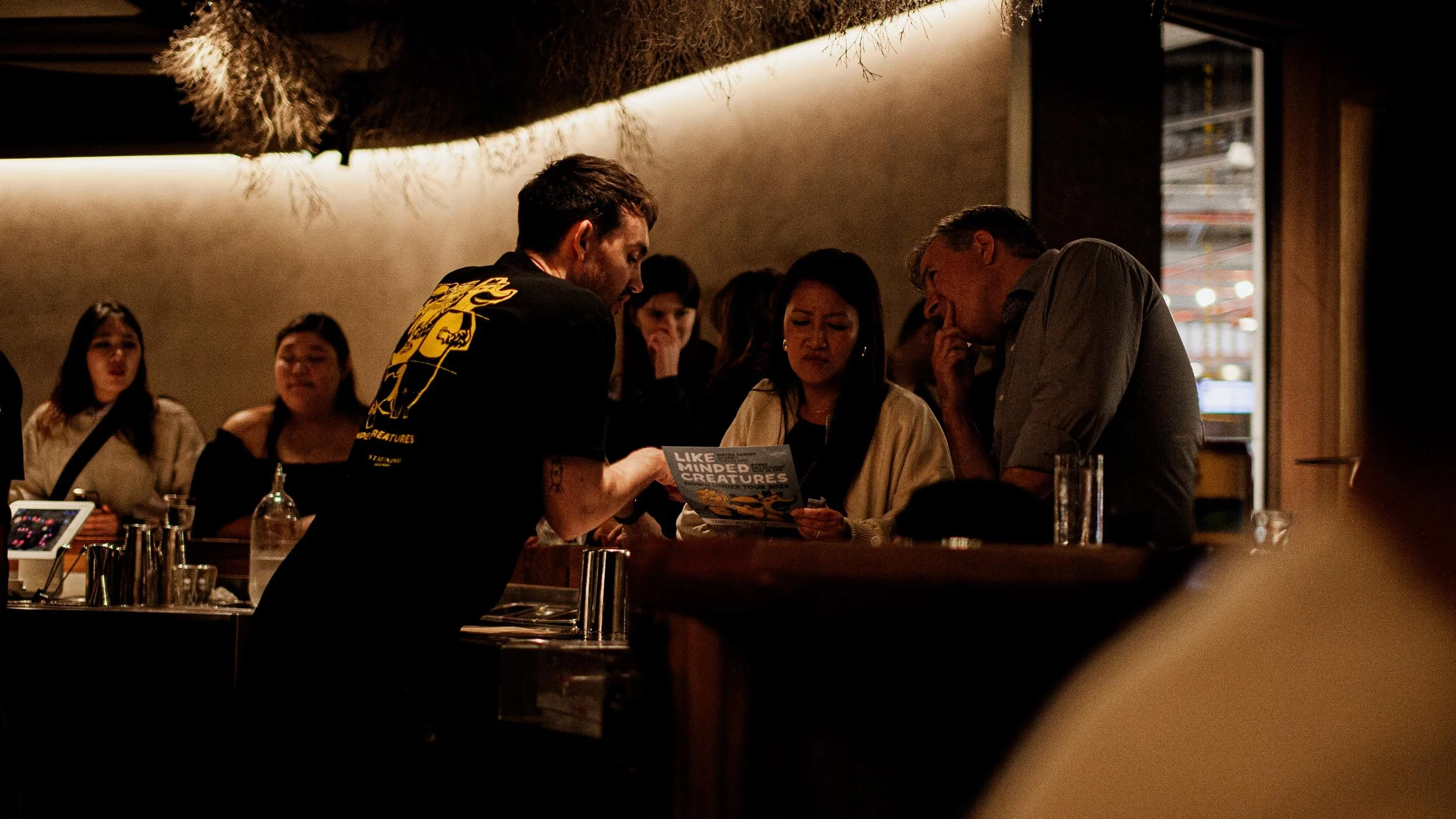 Mid view of bartender Sam Thornhill pointing out something on a Like Minded Creatures menu at the Byrdi bar. Two customers listen avidly.