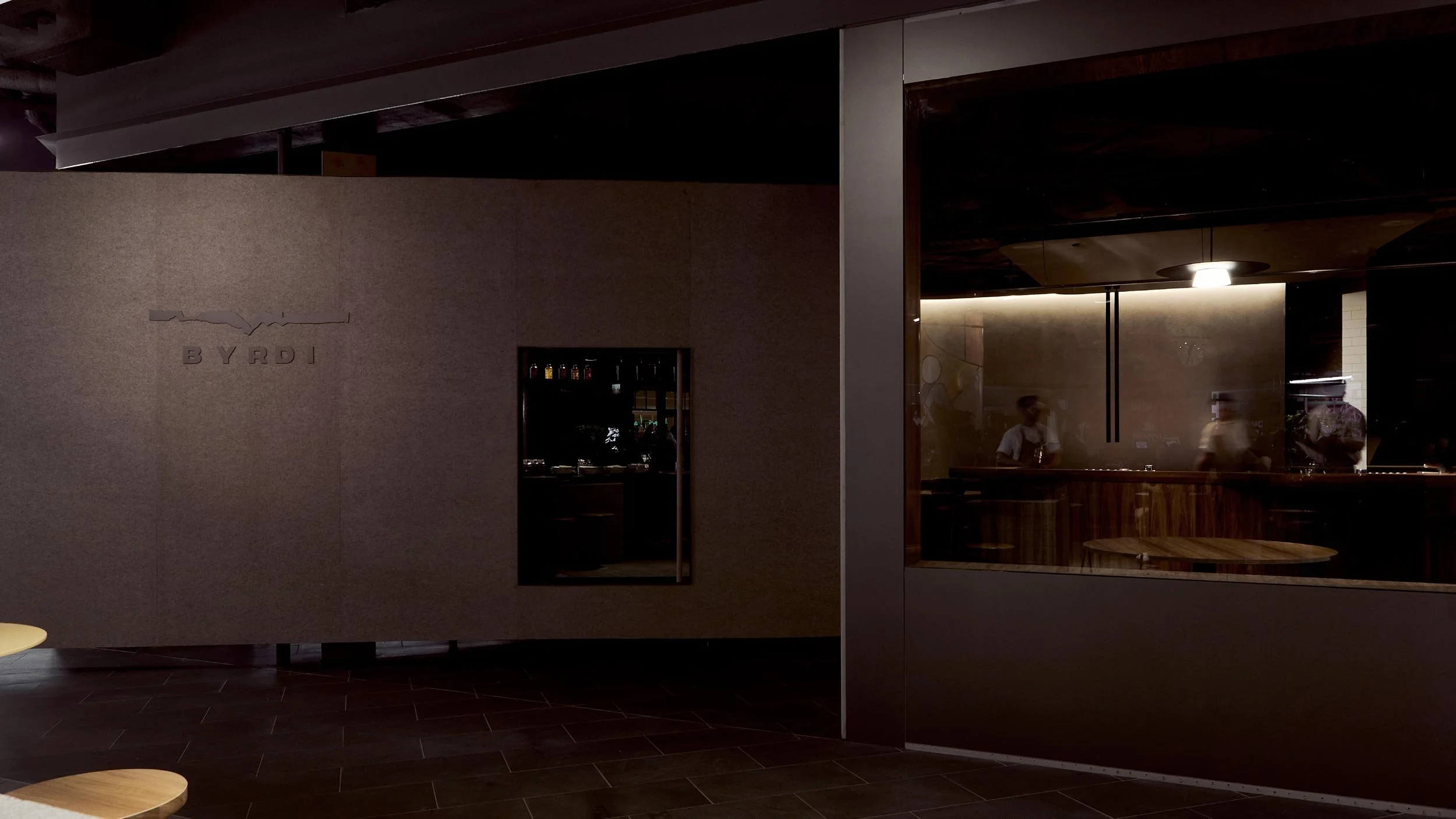 Exterior of Byrdi bar showing logo laser cut out of steel on the oversized pivoting door. Through the window we can see three bartenders working.