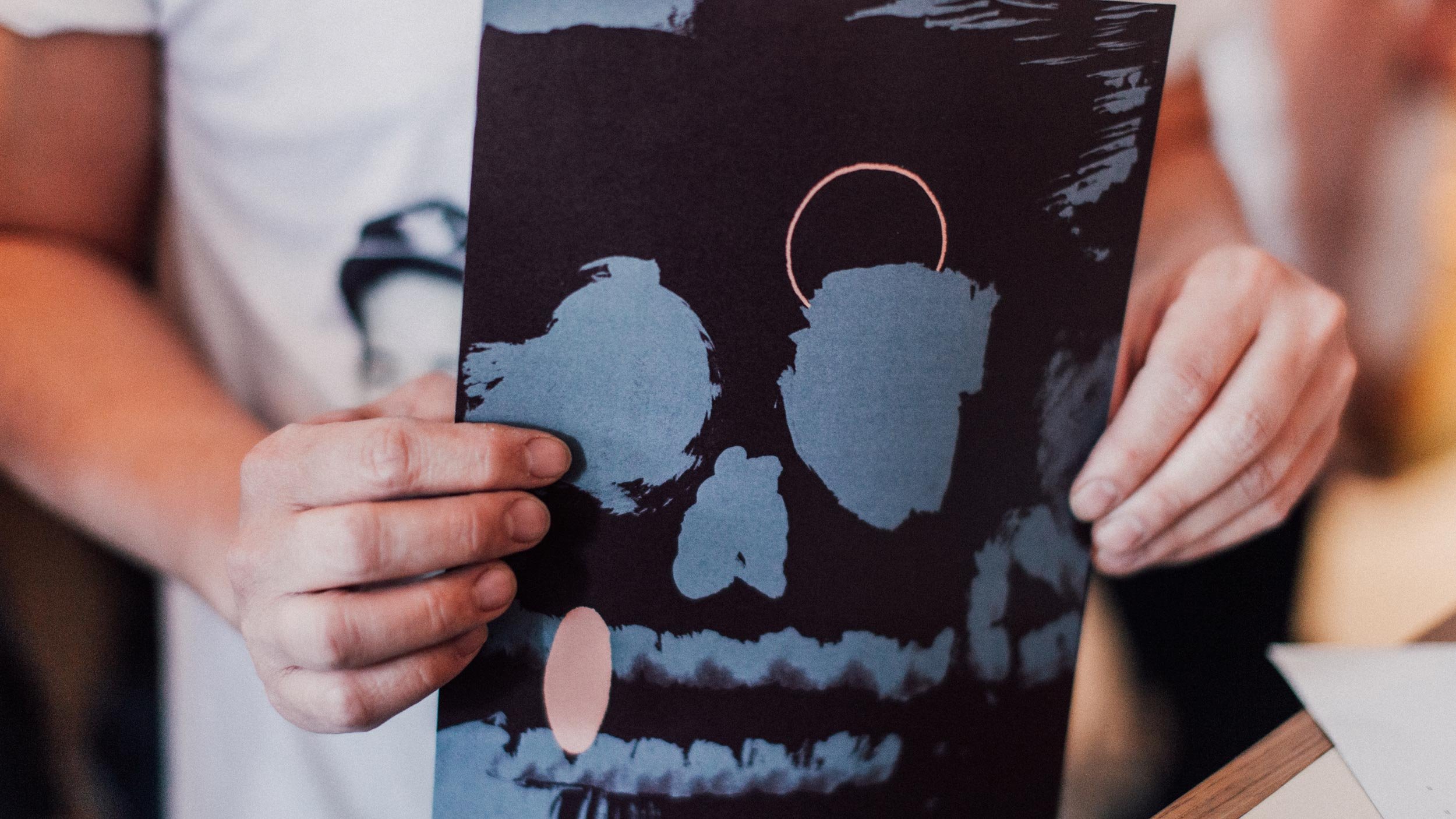 Close-up view of hands holding a Someday Sessions menu. We can see an abstract illustration in maroon and blue on the back.